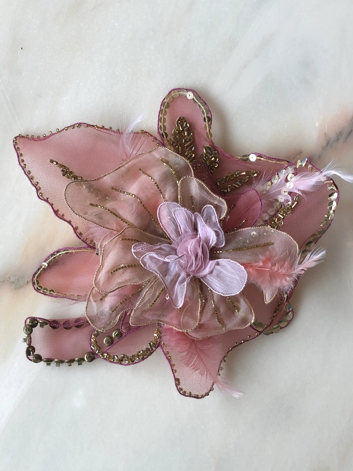 Broochella Alstroemeria Large handmade designer flower brooch in layered blush pink and mauve organza, intricately hand-beaded with gold beads and sequins and finished with delicate feather details, photographed on pale marble with natural grey veining.