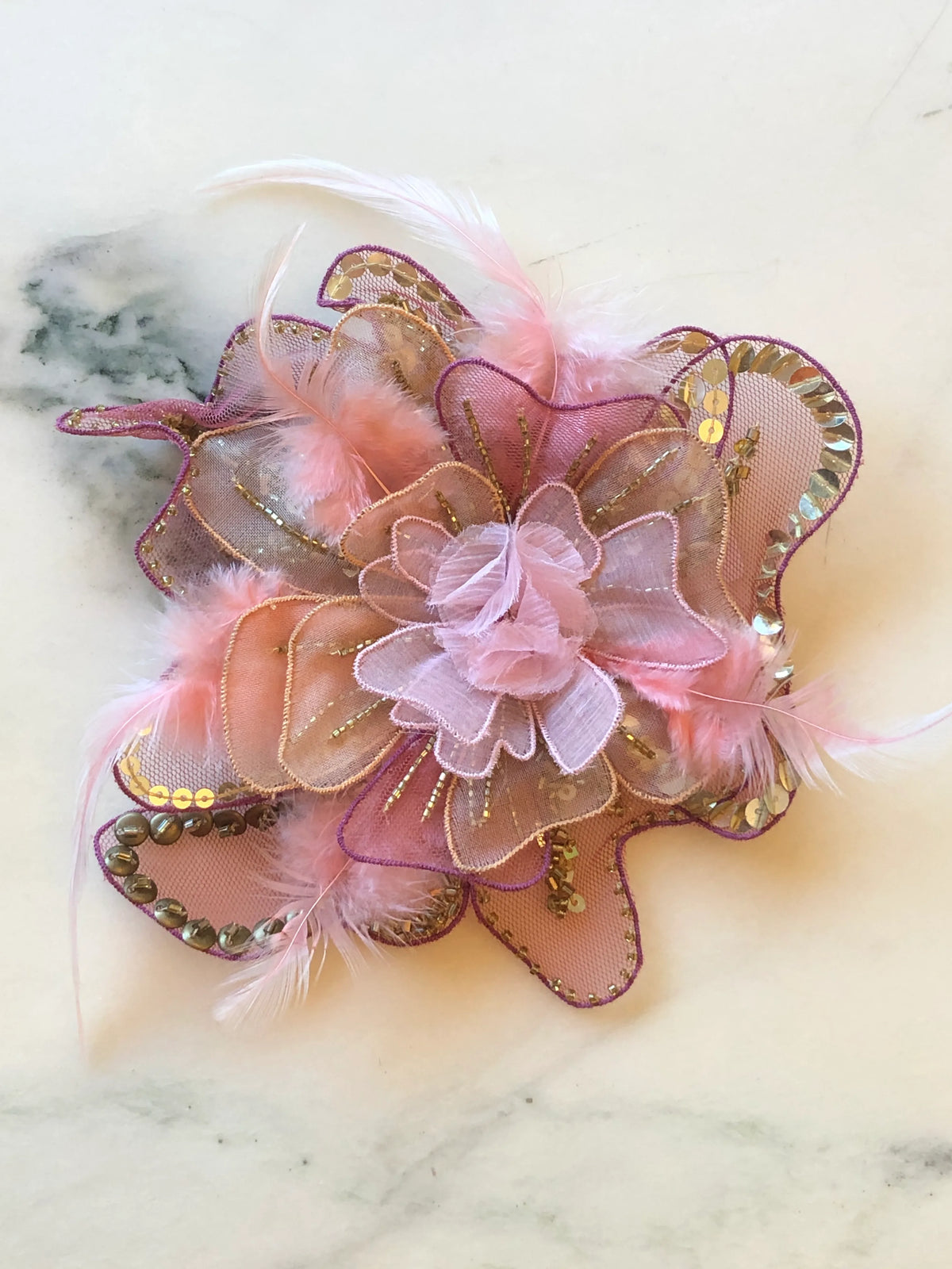 Broochella Alstroemeria Hand-Beaded Flower Brooch - Small in soft pink, featuring layered sheer organza petals embellished with gold sequins and beads, purple edging and delicate pink feather accents on a cream marble background.