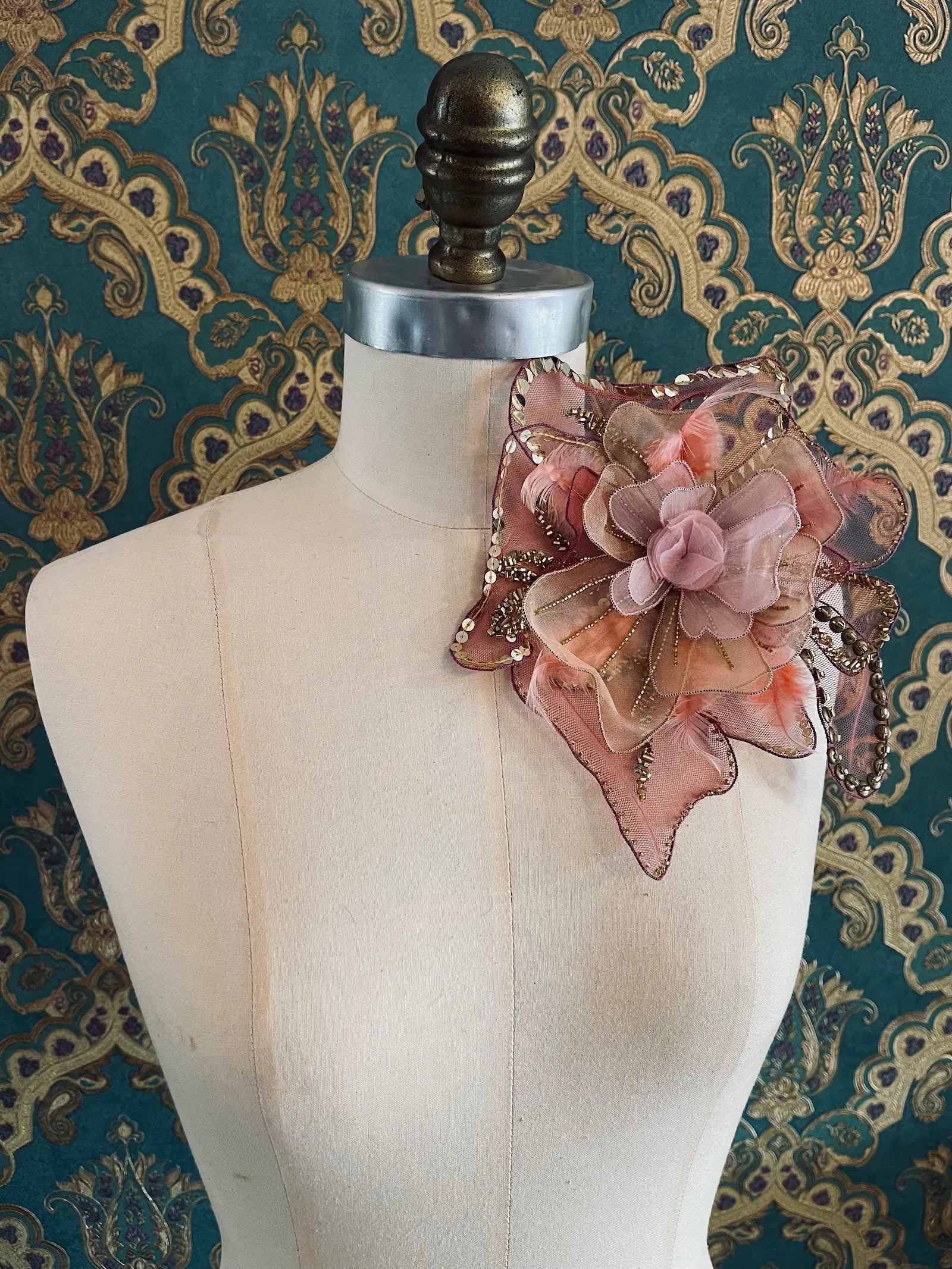 Broochella alstreomeria hand beaded, large, sequinned, flower brooch in pink and gold. It is pinned to a mannequin. The background is green and gold damask wallpaper.