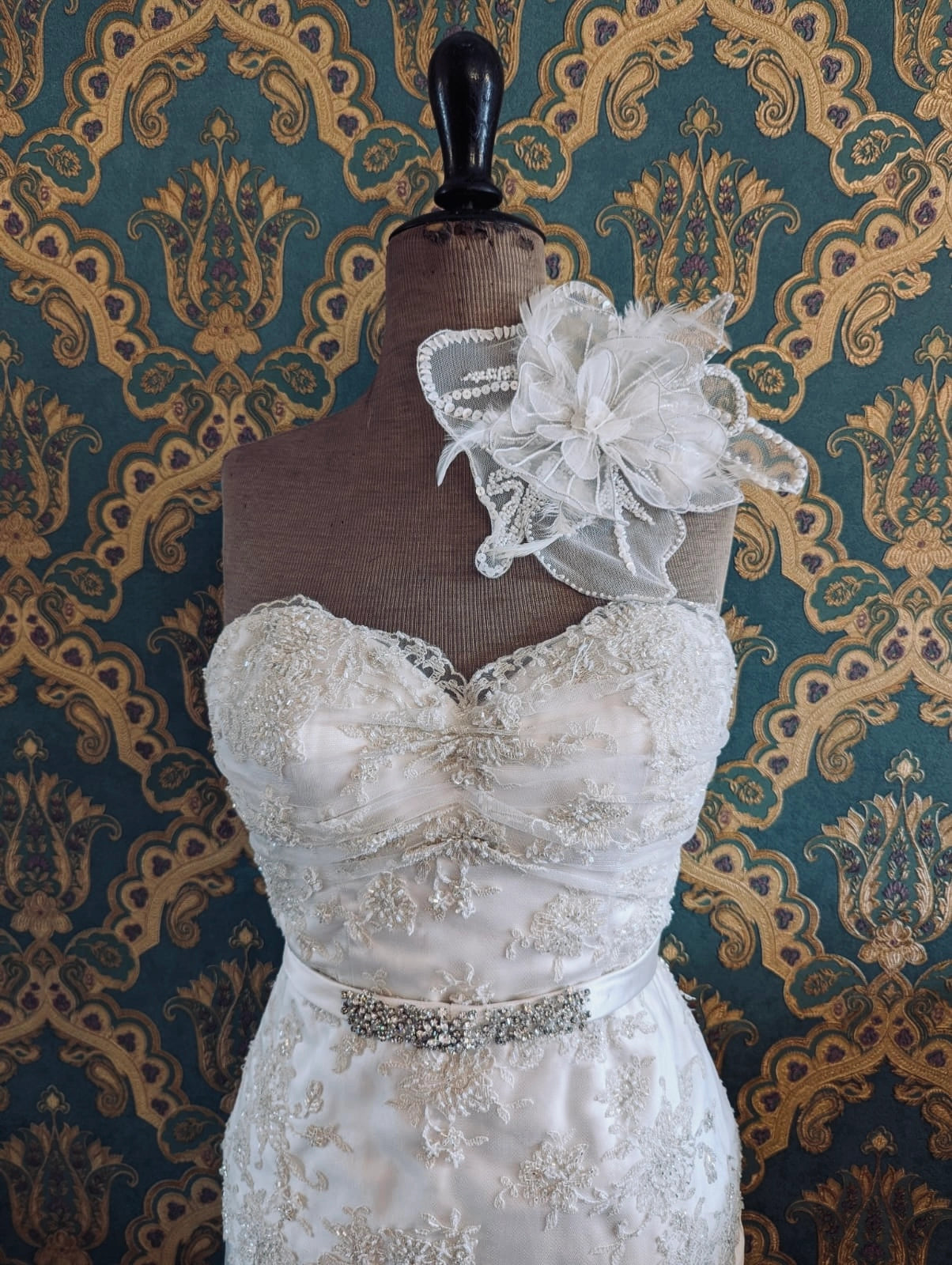 Broochella bridal alstromeria large white handmade beaded and feather flower brooch. Pinned on a mannequin that is wearing a white, beaded strapless wedding dress. The background is green and gold damask wallpaper. 