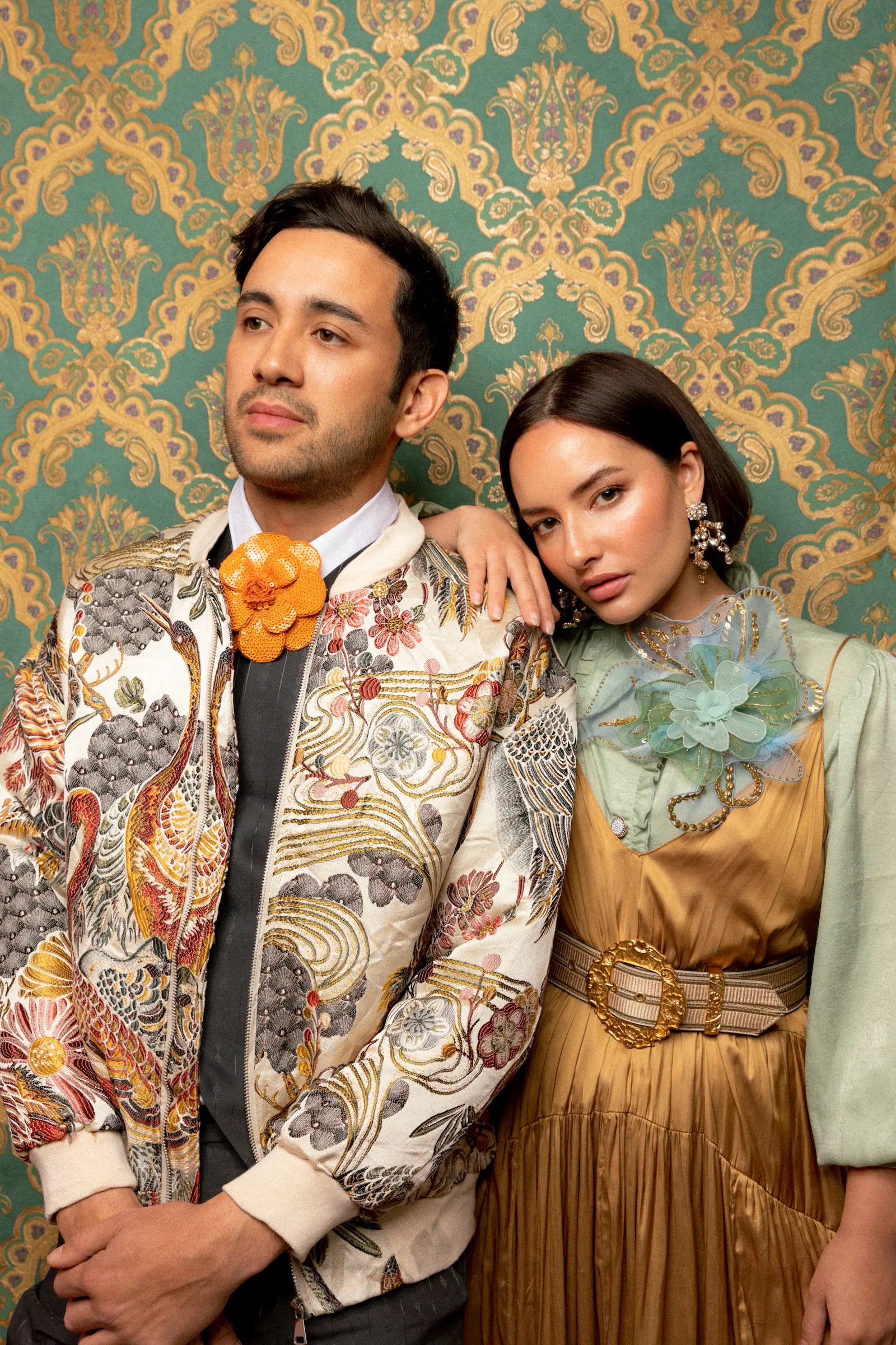 Male and female models with dark hair. She is wearing a gold dress and the Broochella alstromeria large flower brooch He is wearing a printed jacket and the broochella scintillante orange flower brooch. The background is green and gold damask wallpaper