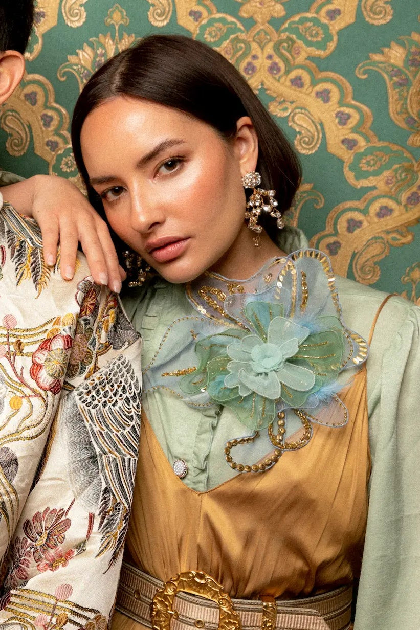 Male and female models with dark hair. She is wearing a gold dress and the Broochella alstromeria large flower brooch He is wearing a printed jacket and the broochella scintillante orange flower brooch. The background is green and gold damask wallpaper