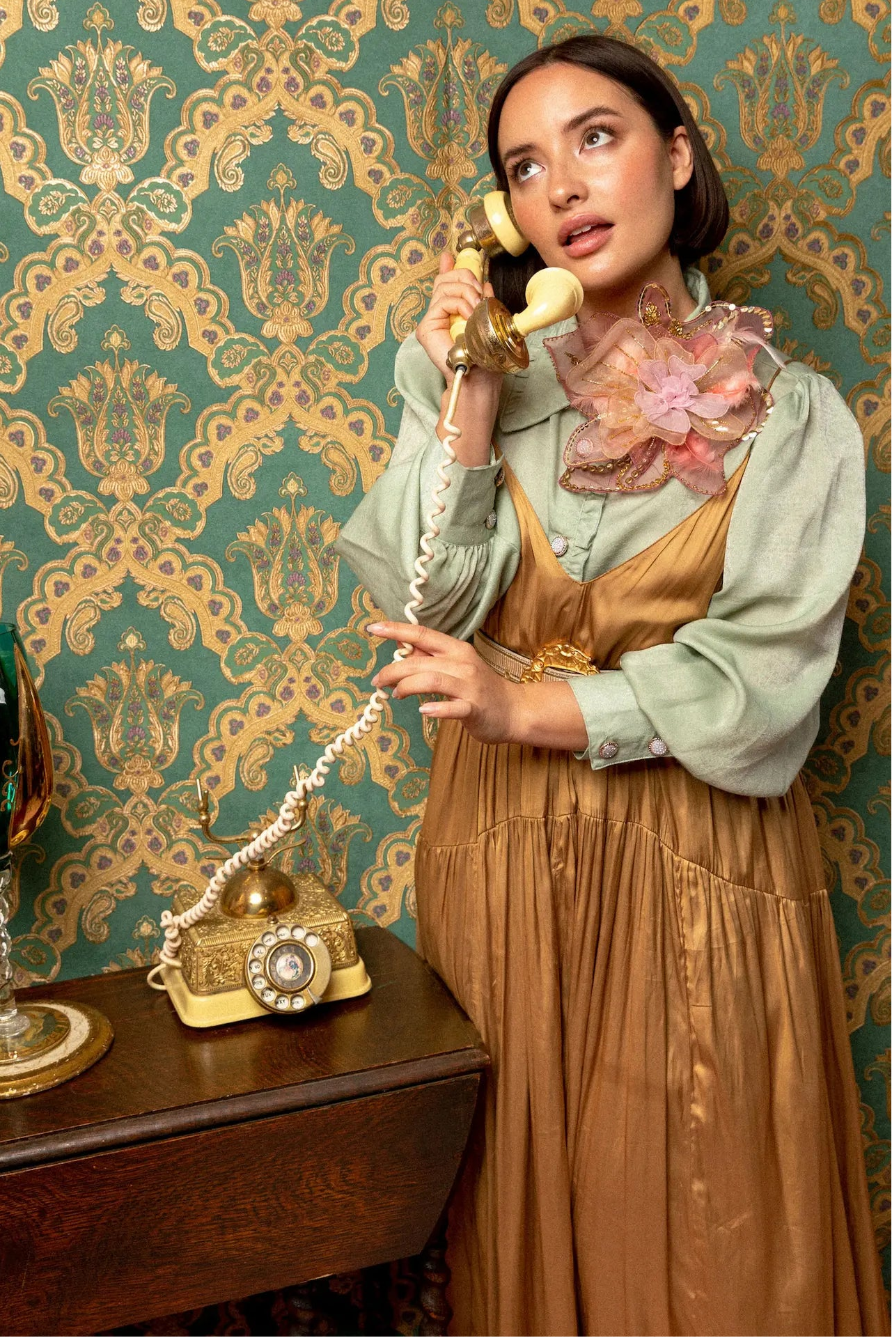 Woman in vintage attire talking on a yellow phone against ornate wallpaper. She is wearing the Broochella alstromeria lage flower brooch in pink and gold