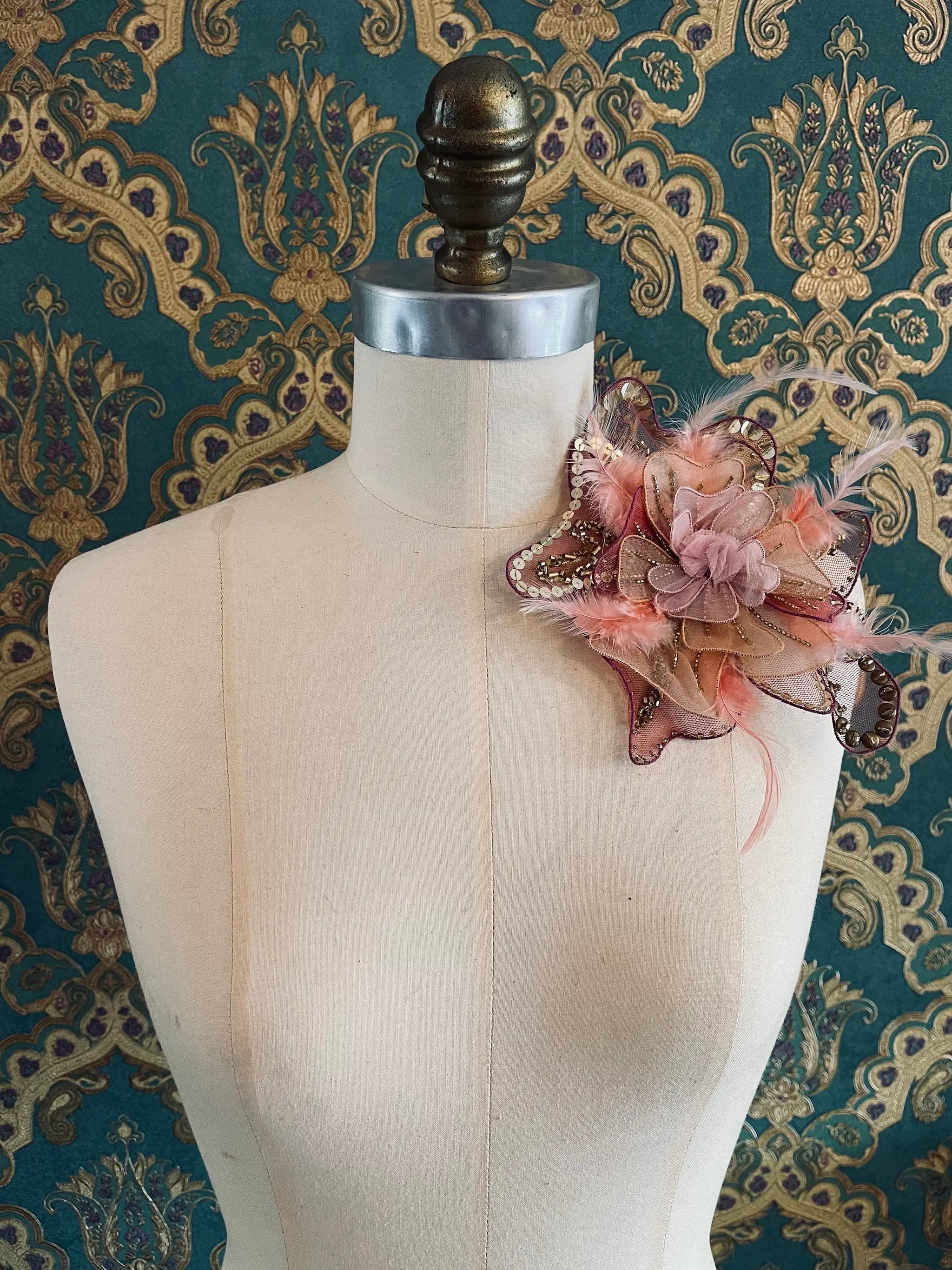 Broochella Alstromeria small, hand beaded designer flower brooch in pink and gold. Pictured on a mannequin. The background is green and gold damask wallpaper