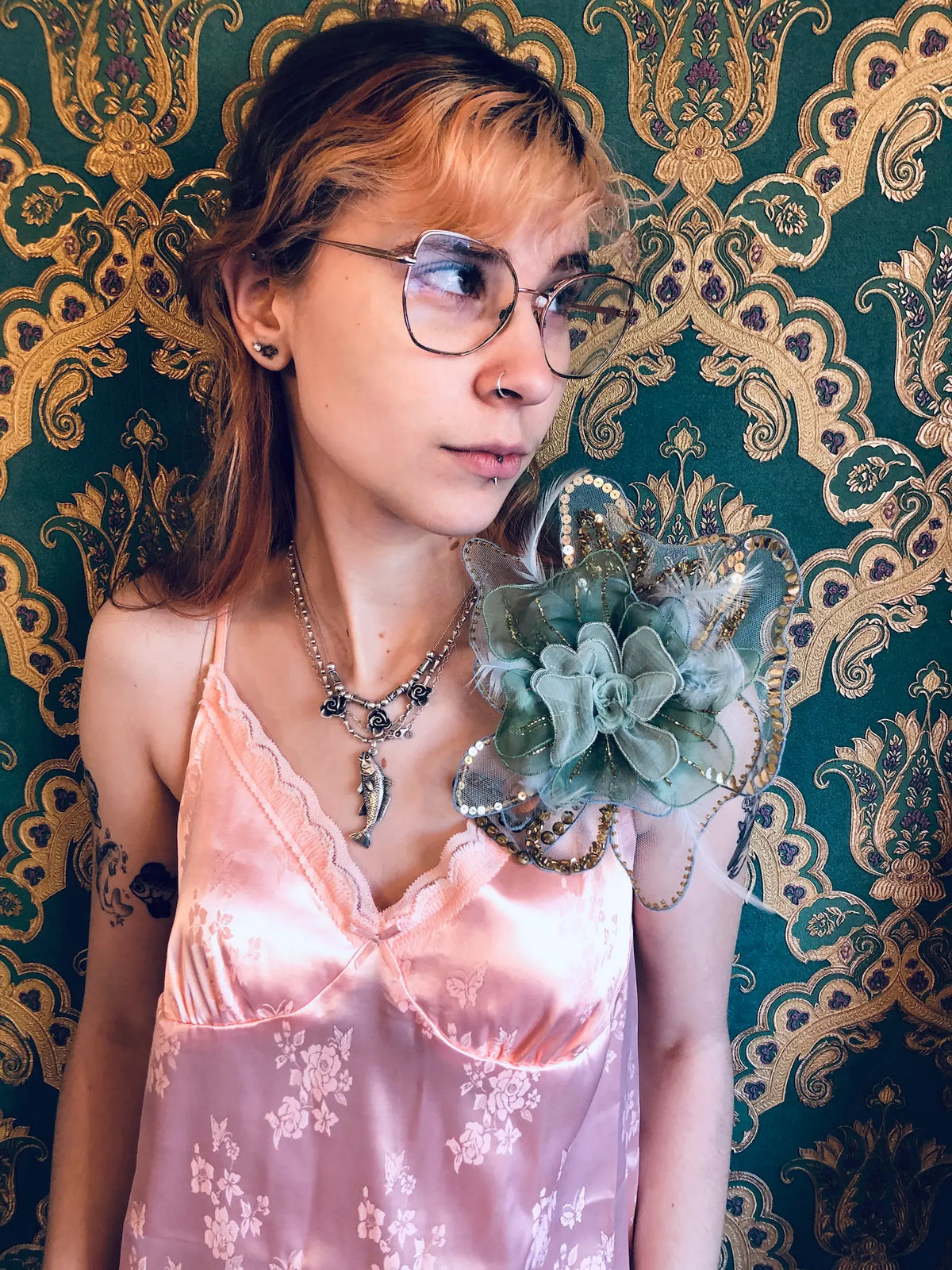 A person with glasses and tattoos poses against a dark green and gold damask patterned wallpaper, wearing a pale pink satin camisole and a Broochella Alstromeria brooch in green/blue.