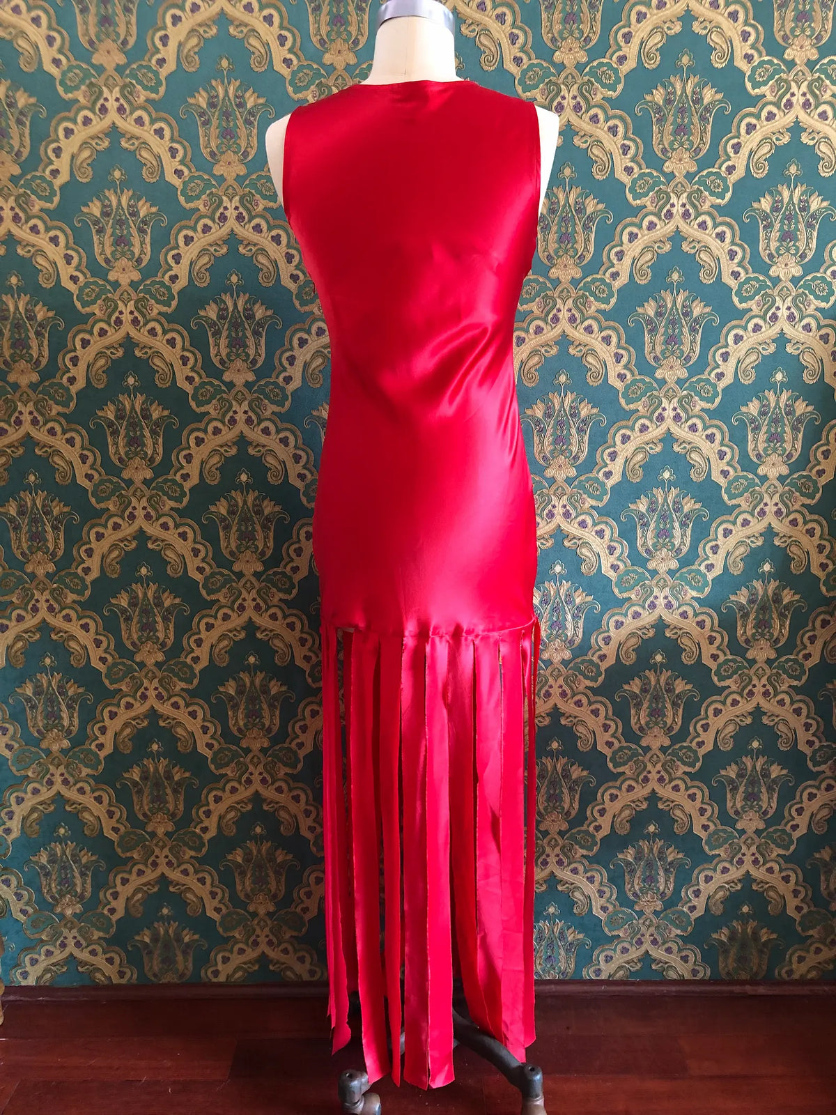 Broochella Amāre scarlet red silk maxi dress with a round neckline, sleeveless bodice and dramatic long panelled fringe, size AU 10. Pictured on a mannequin against green and gold damask wallpaper.