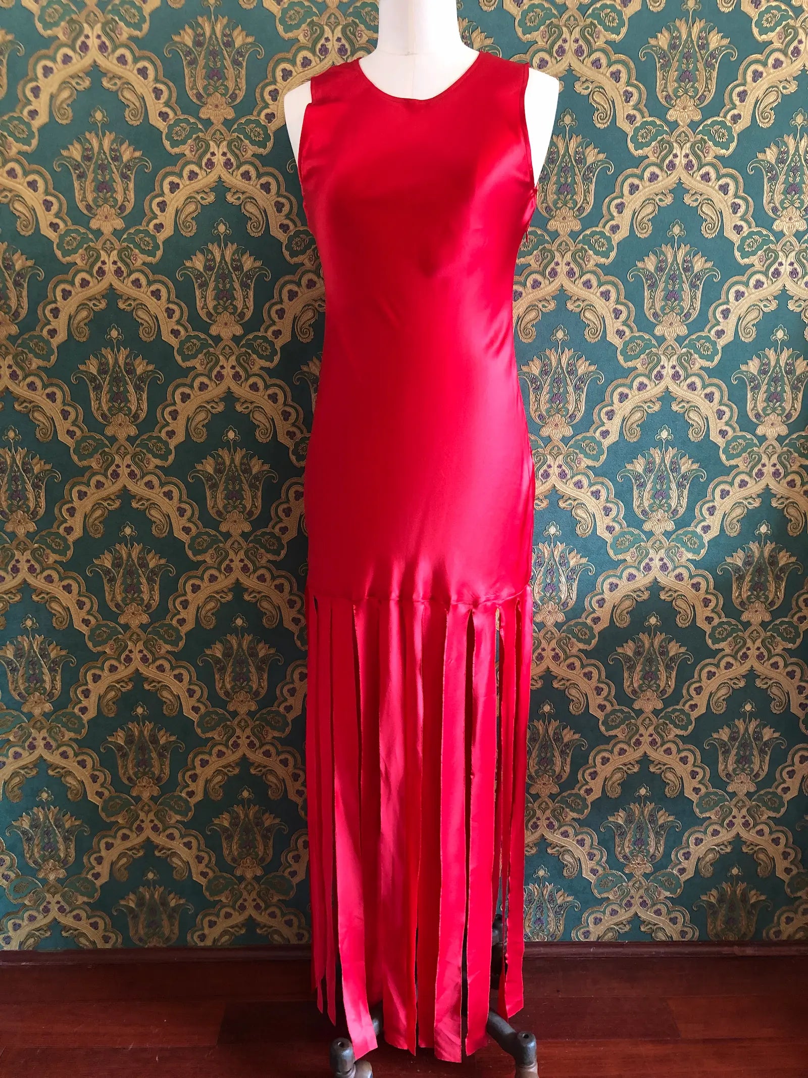 Broochella Amāre scarlet red silk maxi dress with a round neckline, sleeveless bodice and dramatic long panelled fringe, size AU 10. Pictured on a mannequin against green and gold damask wallpaper.