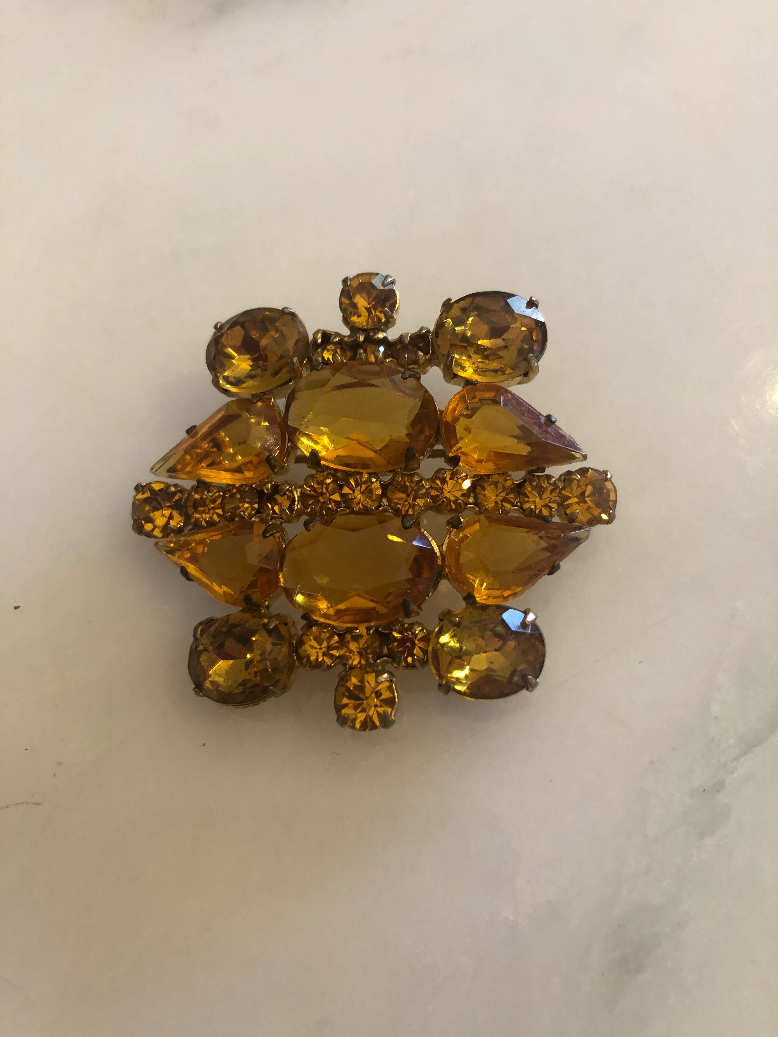A Broochella ambra vintage gold-tone rhinestone brooch featuring multiple faceted amber-yellow crystals arranged in a floral or snowflake-like pattern, resting on a light marble surface.