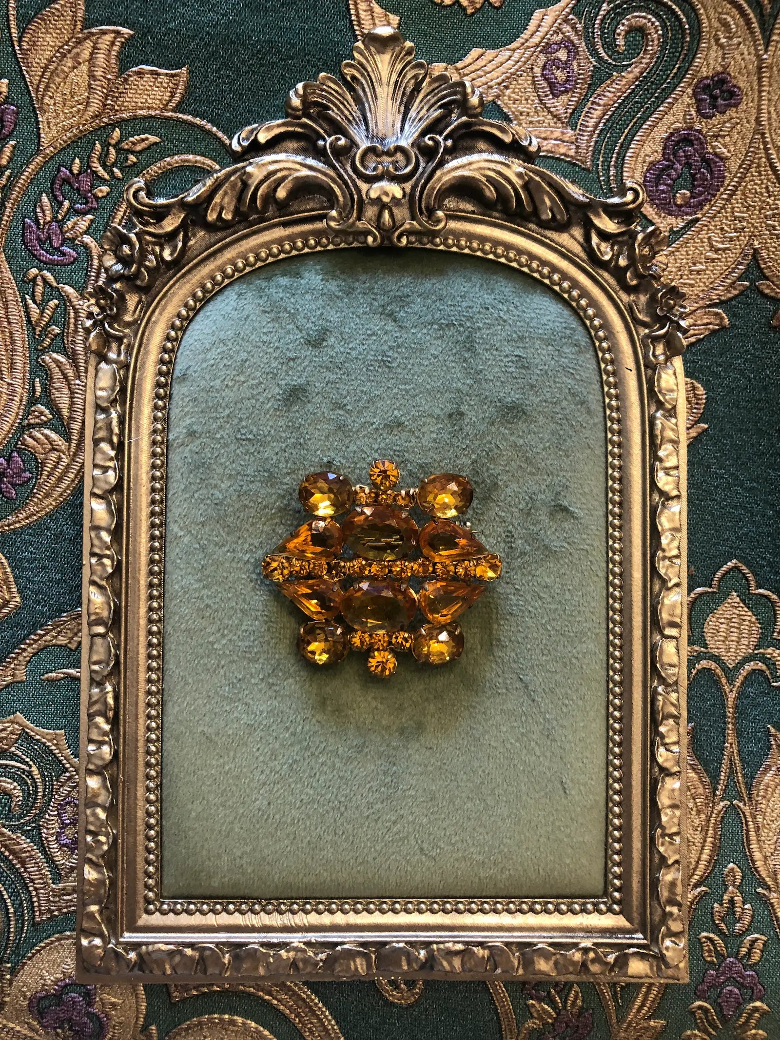 Broochella Vintage ambra crystal gold tone brooch. It is pinned on a green velvet tray with a gold frame. The background is green and gold damask wallpaper. 