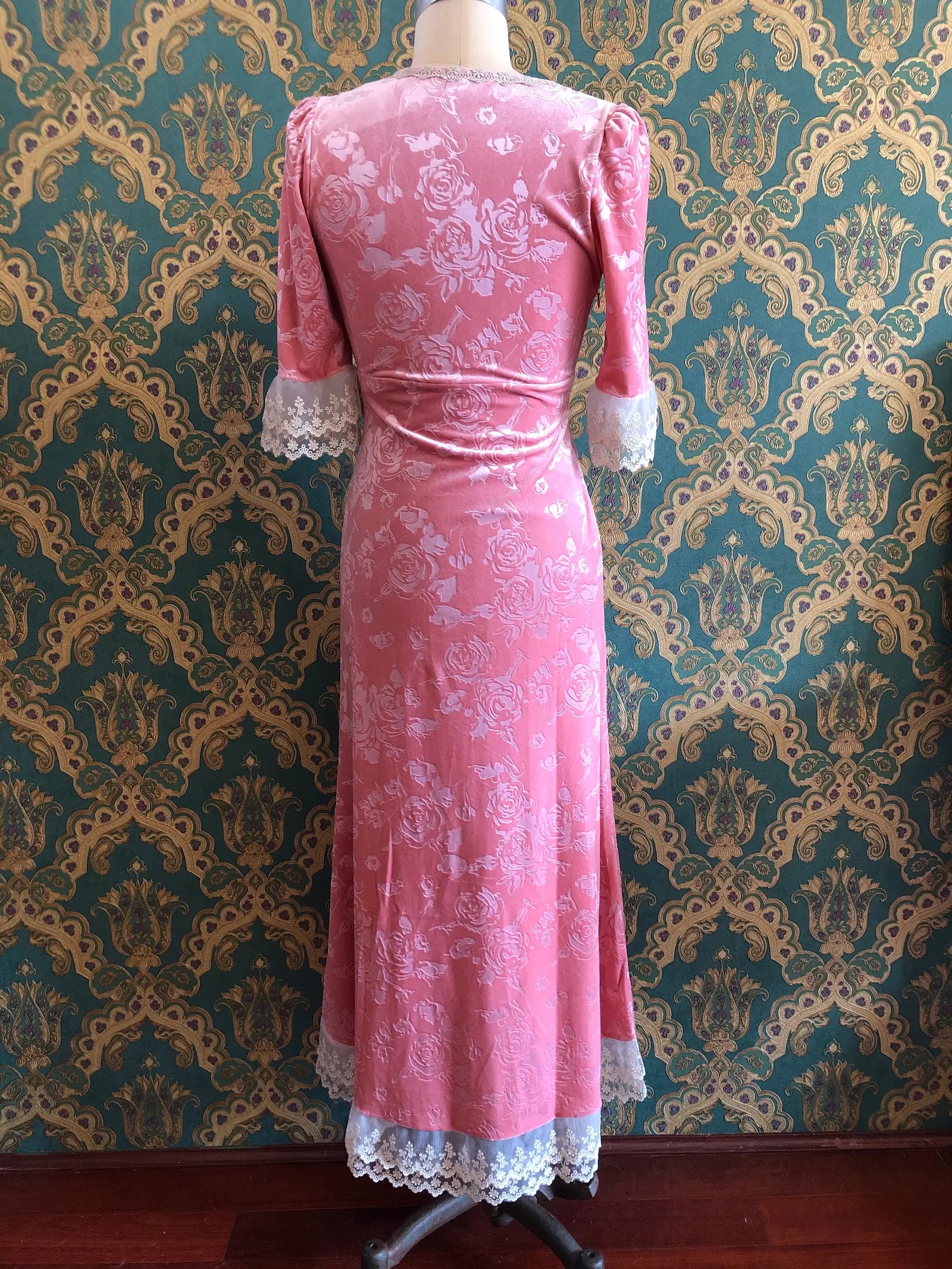 The back view of Broochella American Heiress Gwyneth maxi dress in Dusty Rose Pink with embossed floral velvet, a deep V-neckline and ivory lace trim, size Medium. Pictured on a mannequin against green and gold damask wallpaper.