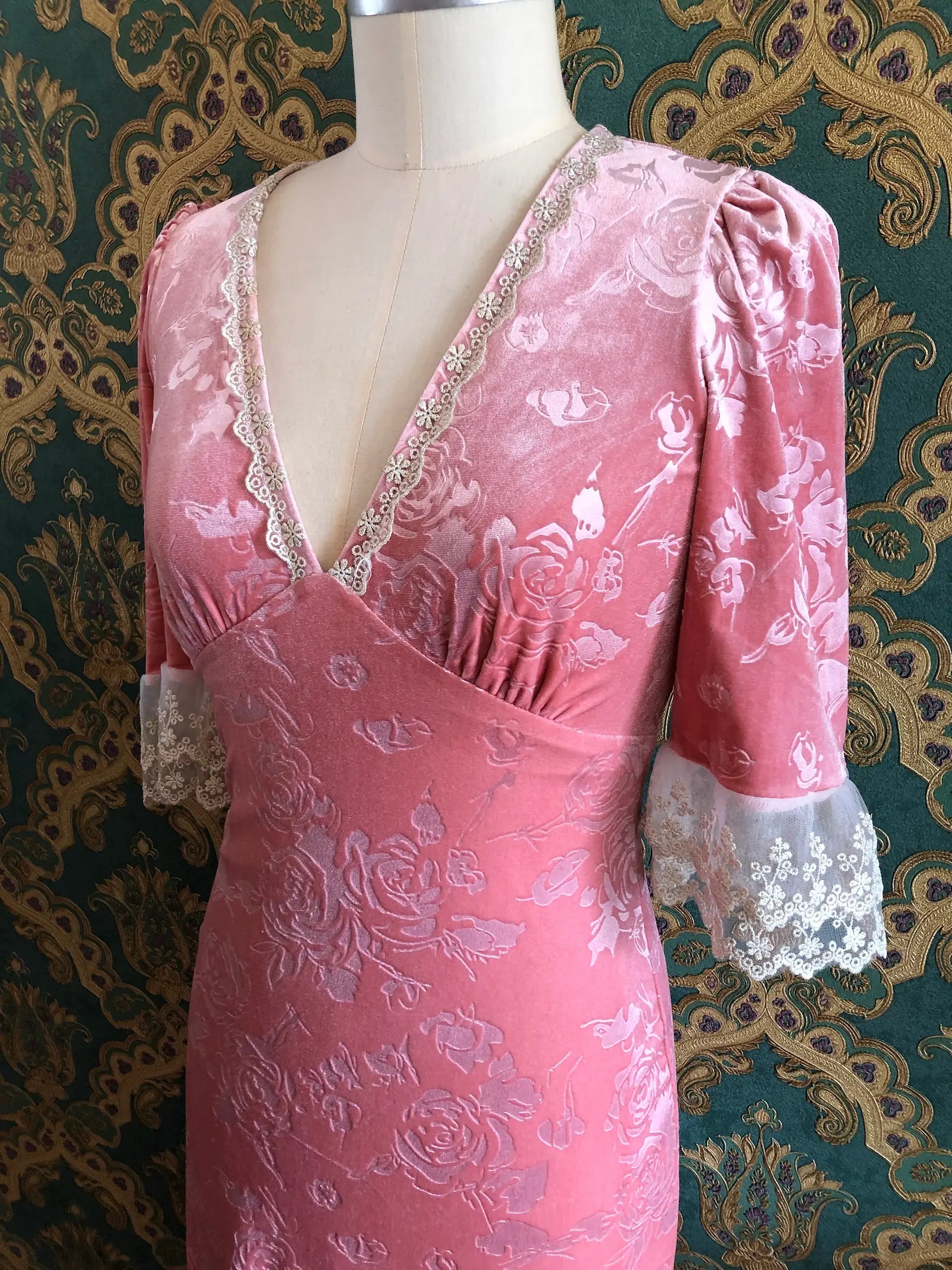 Broochella American Heiress Gwyneth maxi dress in Dusty Rose Pink with embossed floral velvet, a deep V-neckline and ivory lace trim, size Medium. Pictured on a mannequin against green and gold damask wallpaper.