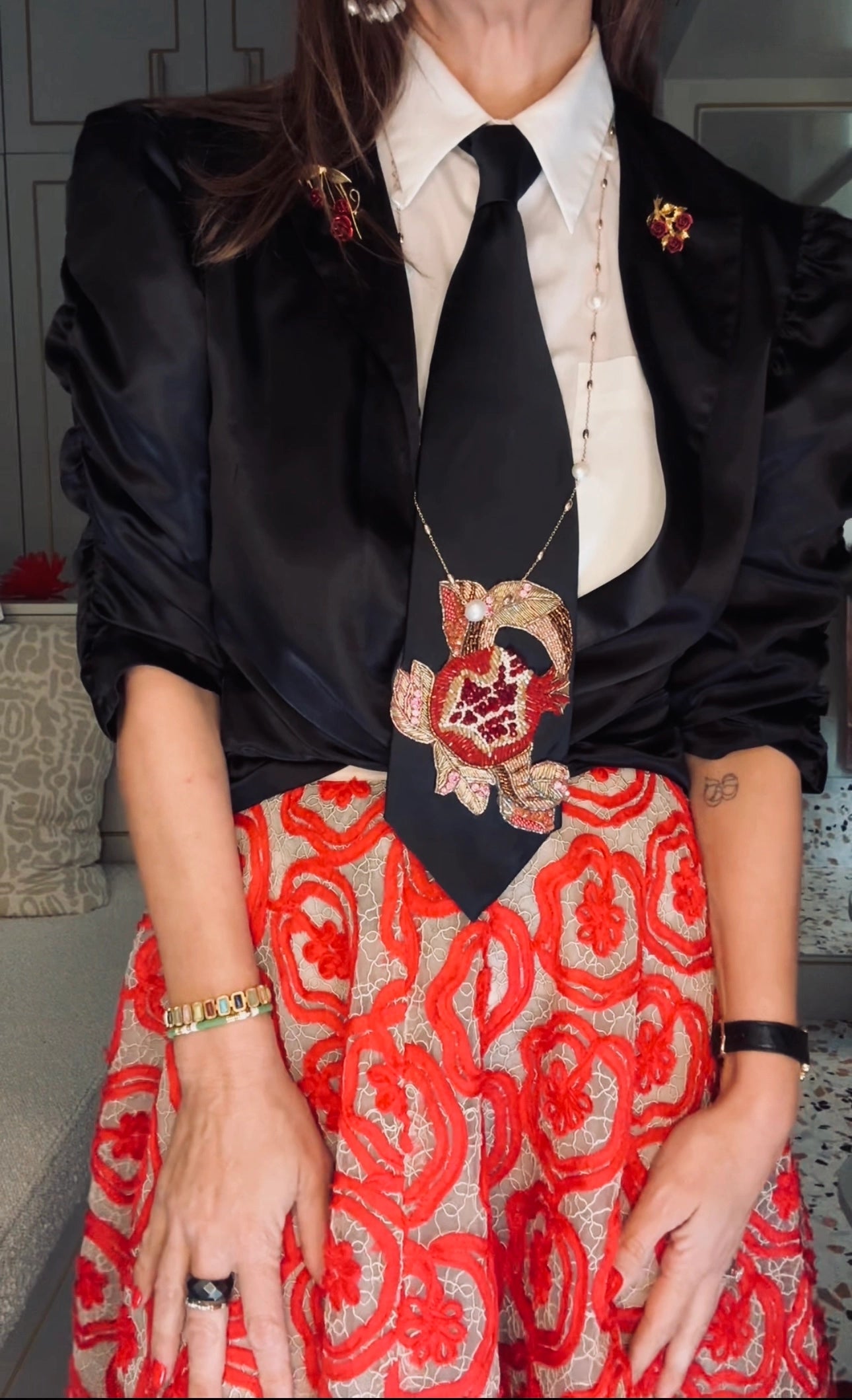 Broochella Amo cravatte embroidered tie. It is pictured on a model in a black jacket and a red skirt. The tie is black has a pomegranate beaded motif.