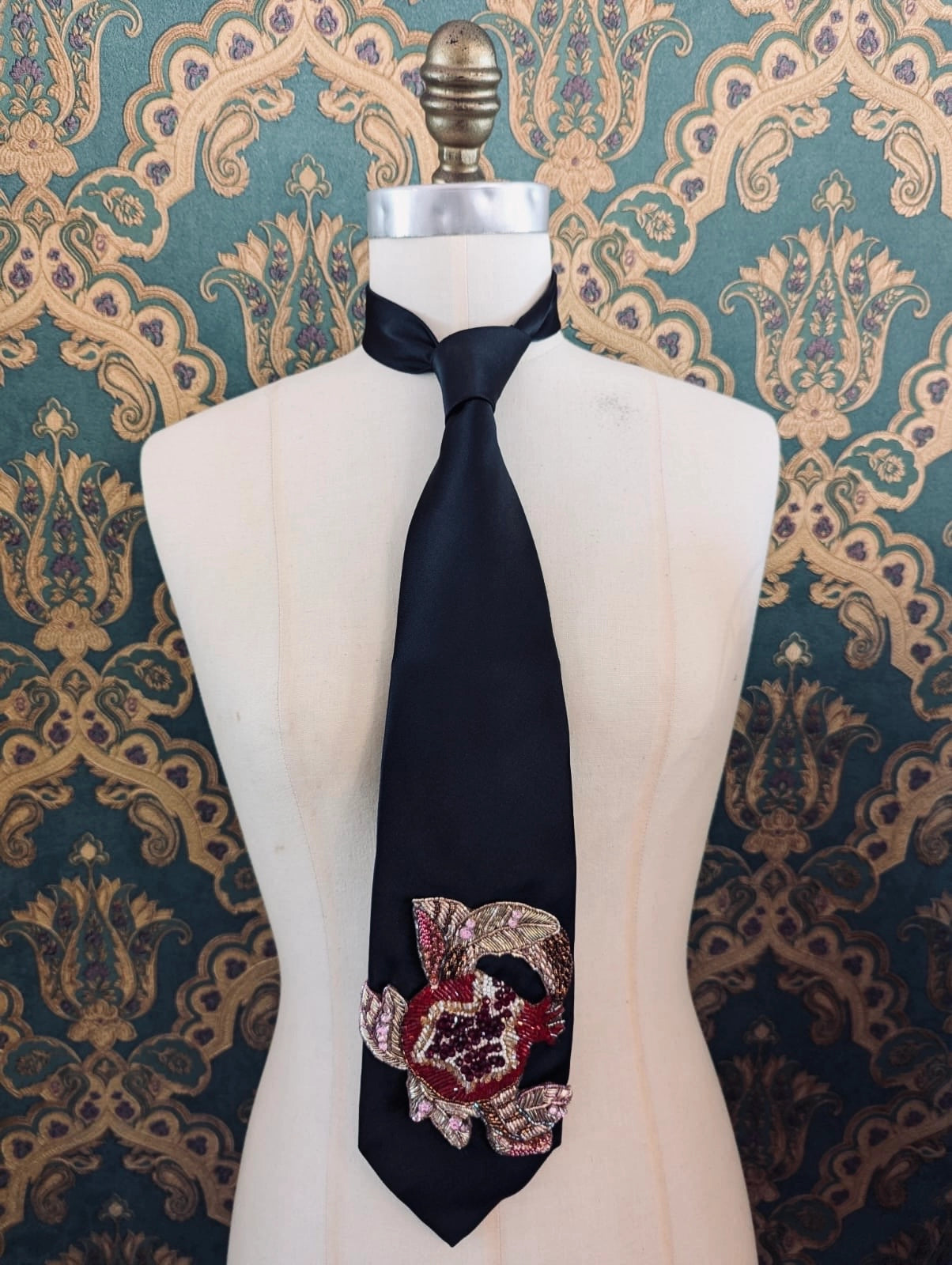 Broochellal amo le cravatta neck tie in black. It has a hand  embroidered pomegranate motif on the front. Pictured on a mannequin. The background is green and gold damask wallpaper.  