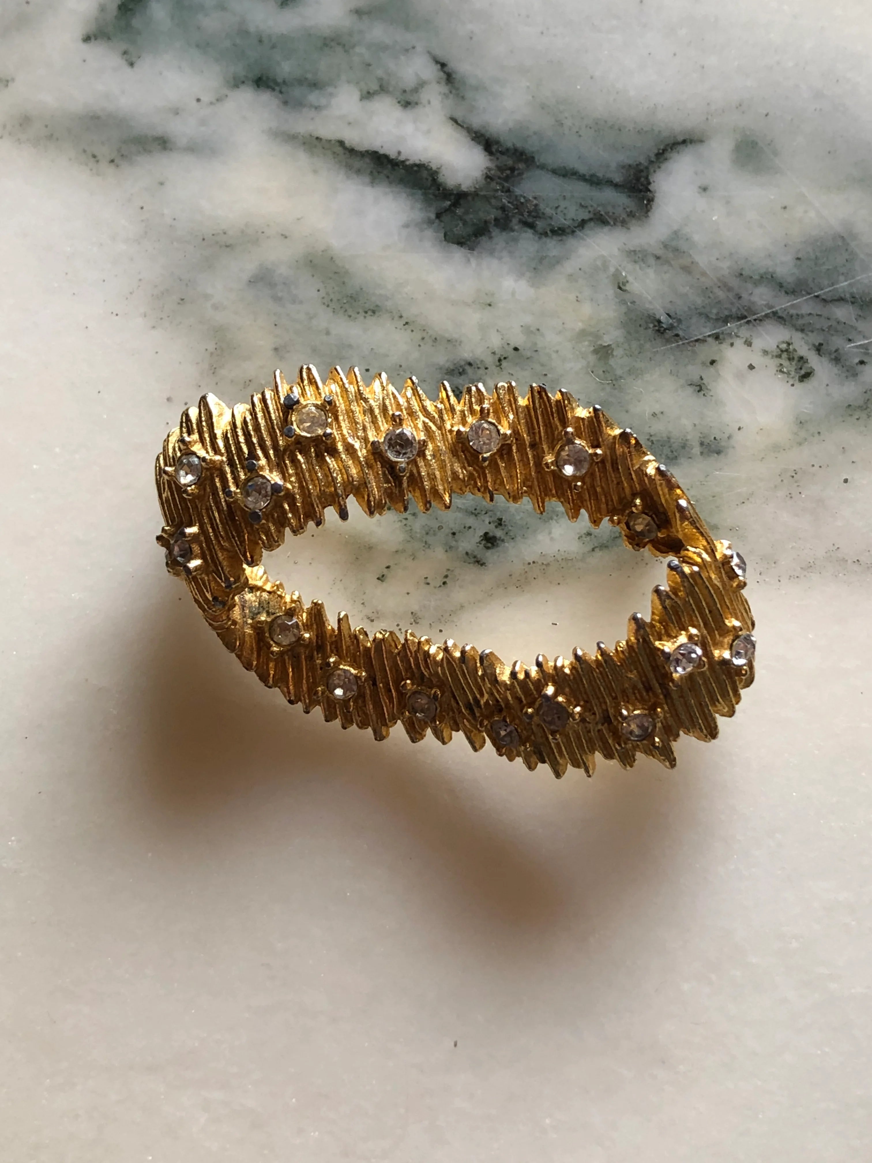 Broochella Anello di Luce vintage gold-tone oval brooch with textured overlapping forms and clear crystal accents. The background is white marble.