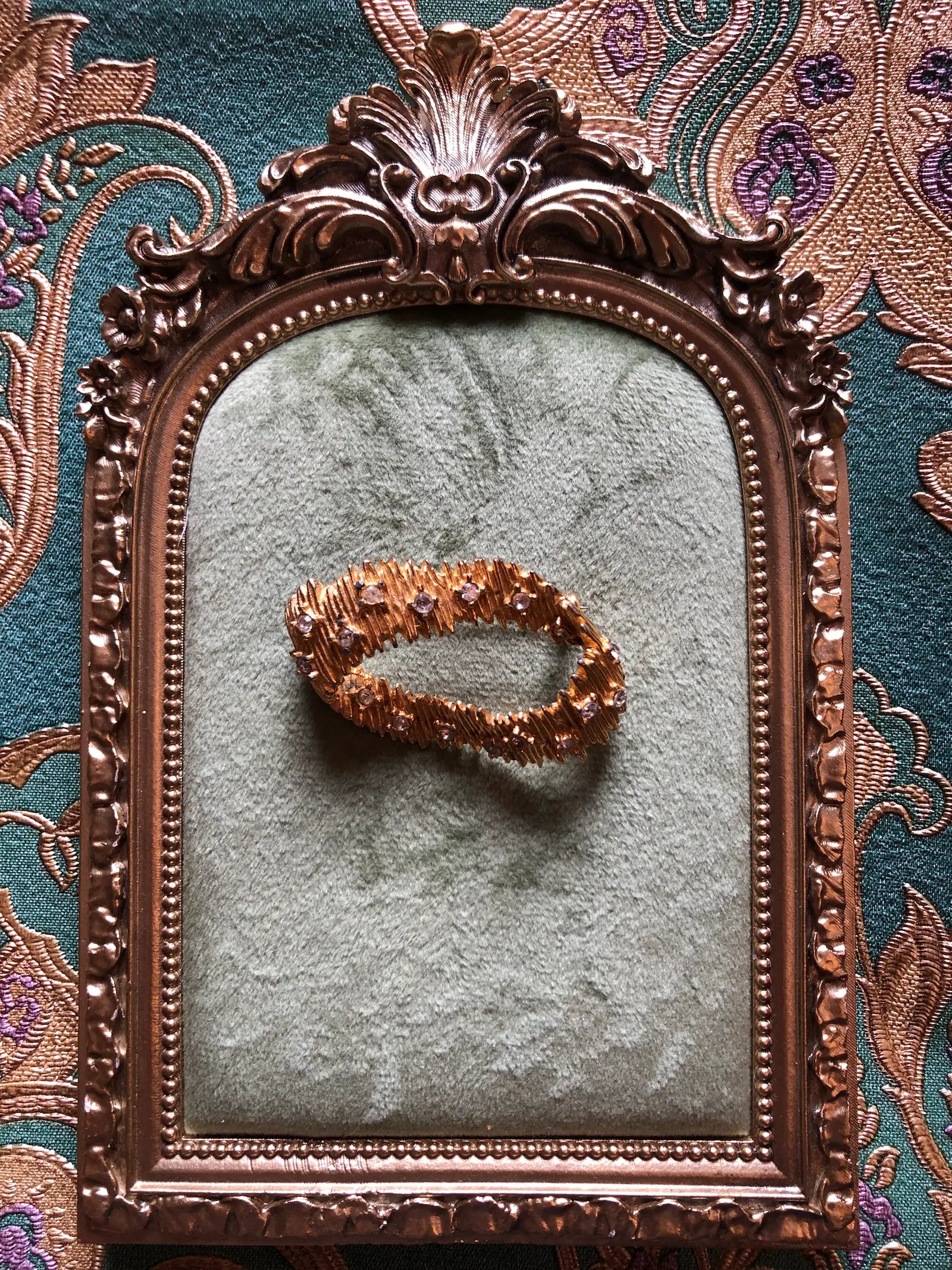 Broochella Anello di Luce vintage gold-tone oval brooch with textured overlapping forms and clear crystal accents. It is displayed on a vintage-style ornate mirror with a green velvet surface, set against a patterned teal and gold fabric background.