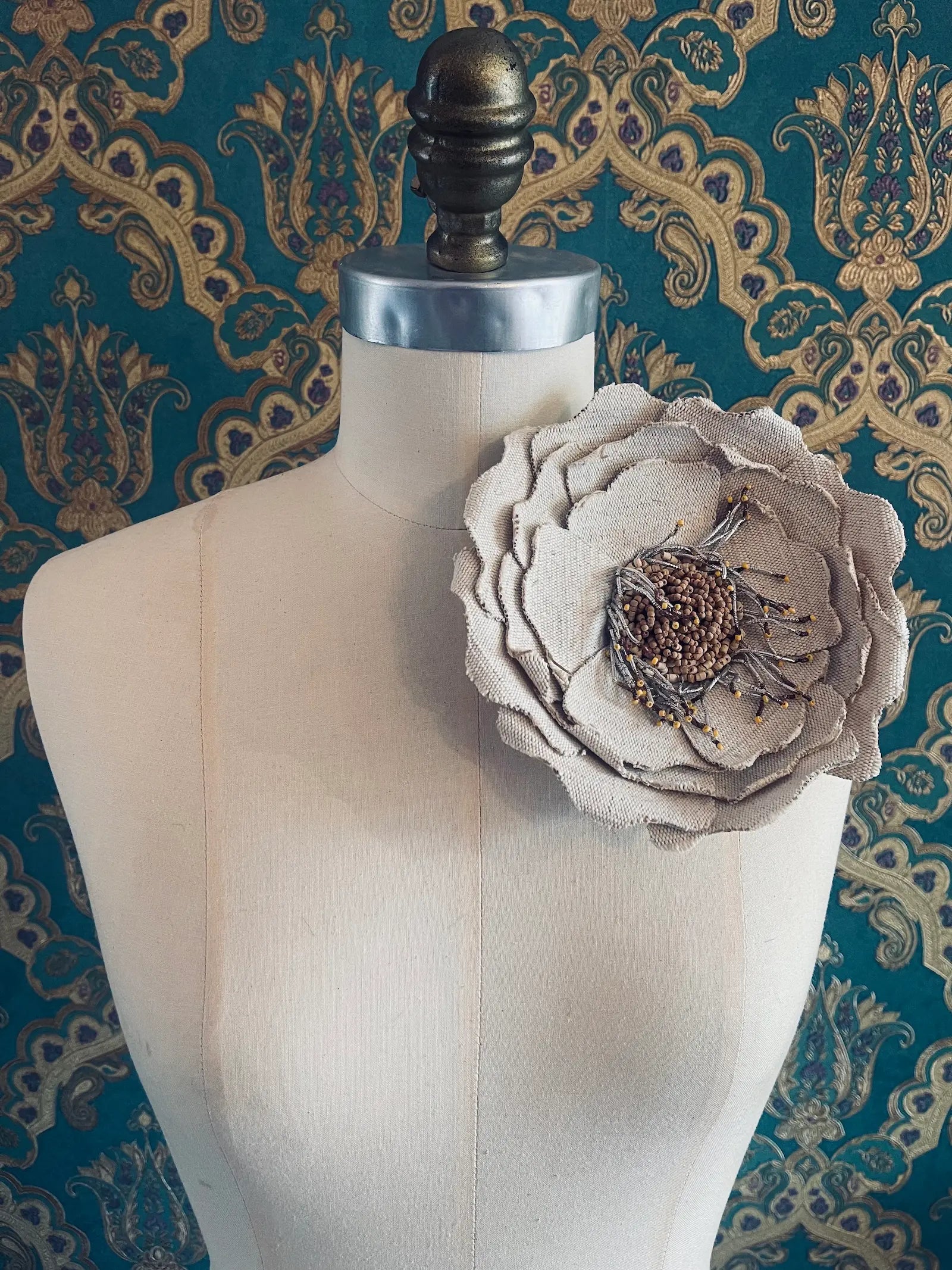 Broochella Anemone large handmade jute fabric flower brooch in beige. Styled on a mannequin. The background is green and gold damask wallpaper