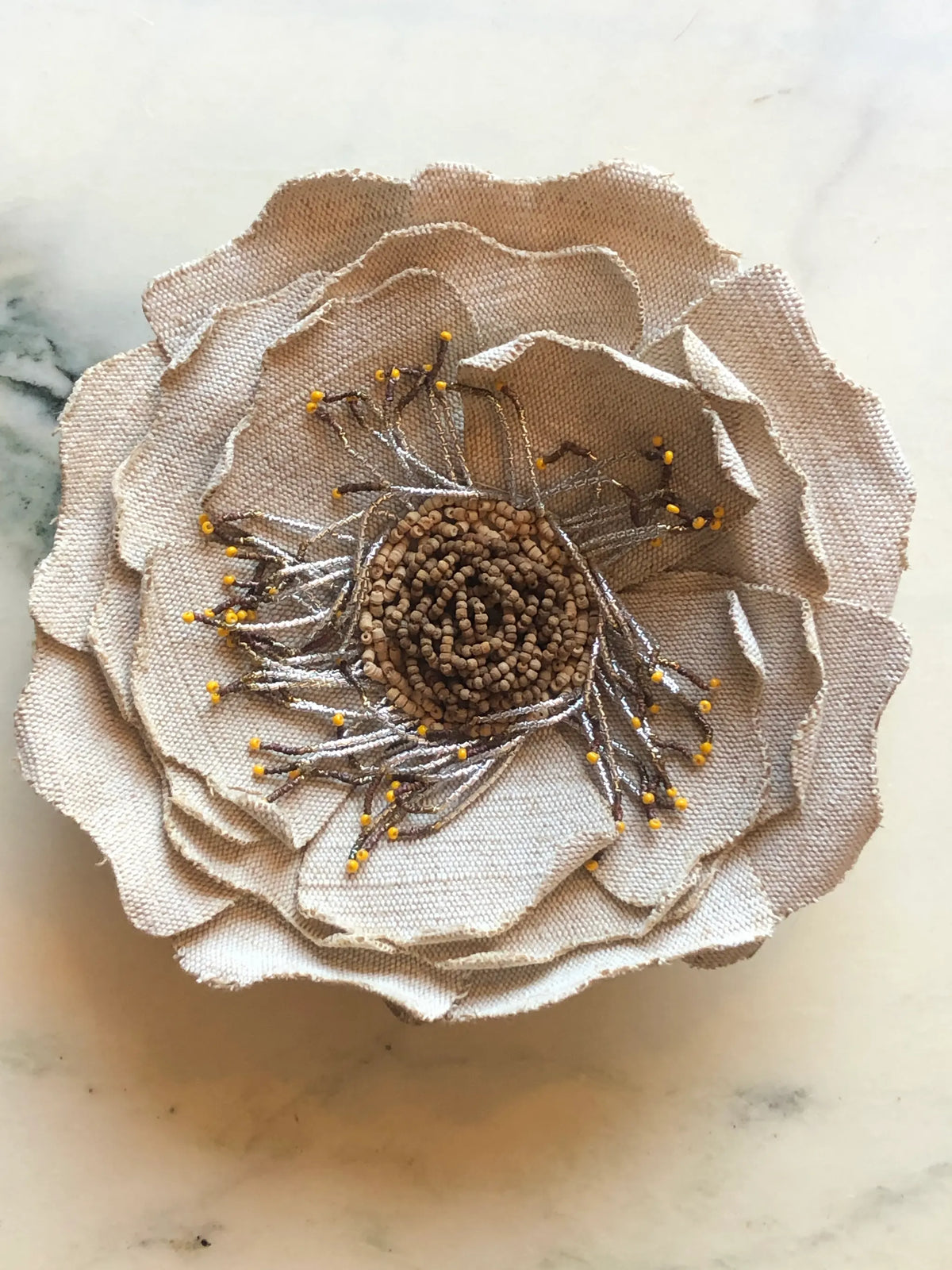 Broochella Anemone Flower Brooch Pin in natural beige, featuring sculptural layered fabric petals with a hand-beaded centre and delicate metallic stamen detailing on a cream marble background.