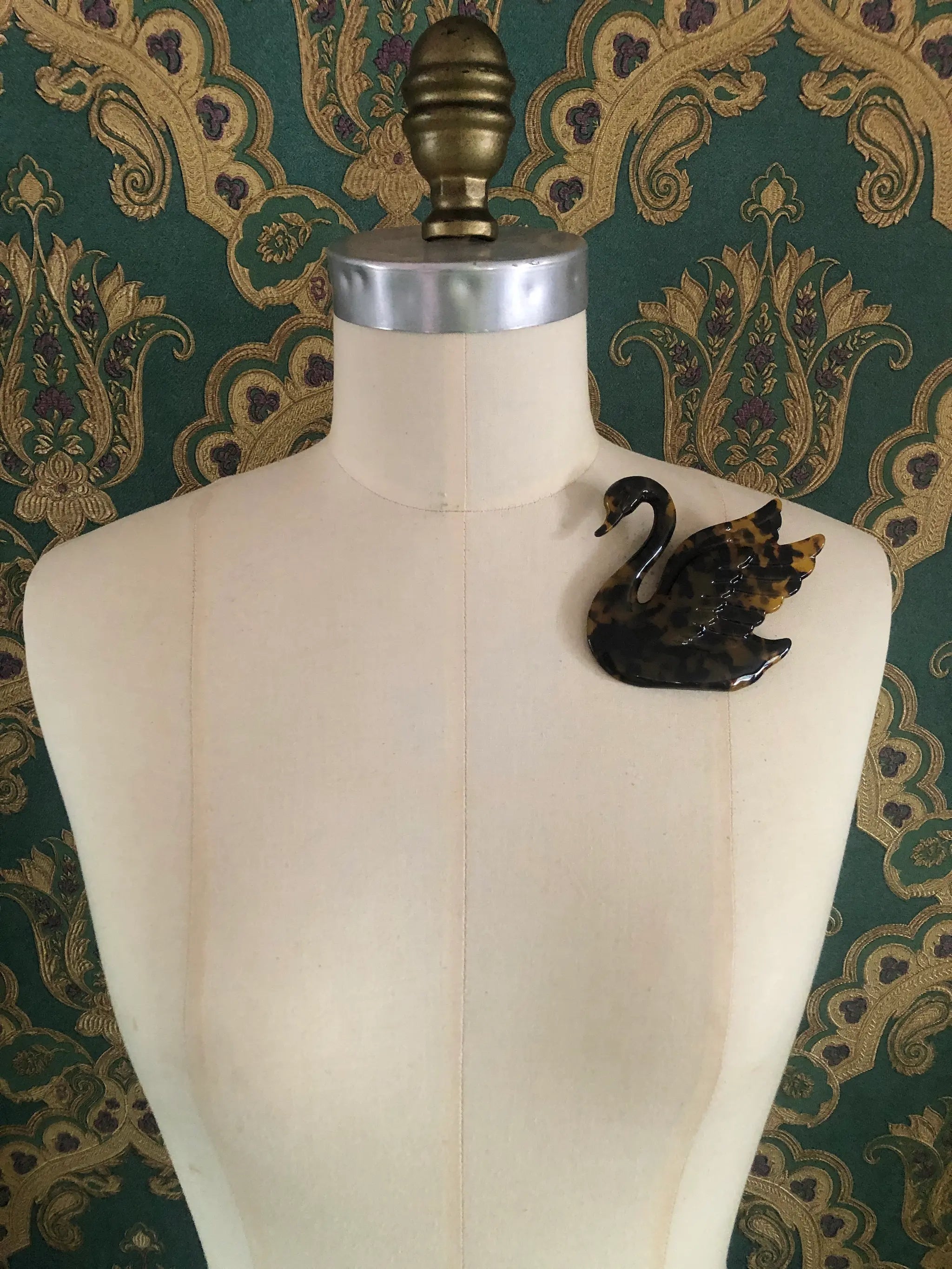 Broochella Animalia brown, figural  hand crafted, cellulose acetate swan brooch in brown tortoiseshell. It is pinned to a mannequin The background is green and gold damask wallpaper.