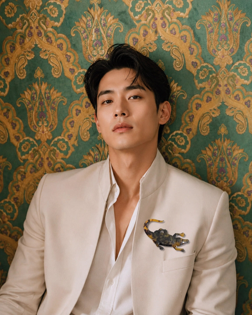 A man wearing a light beige suit jacket and white shirt, leaning against a green damask wallpaper background. He has a Broochella Men's Animalia Tortoiseshell crocodile brooch on his pocket.