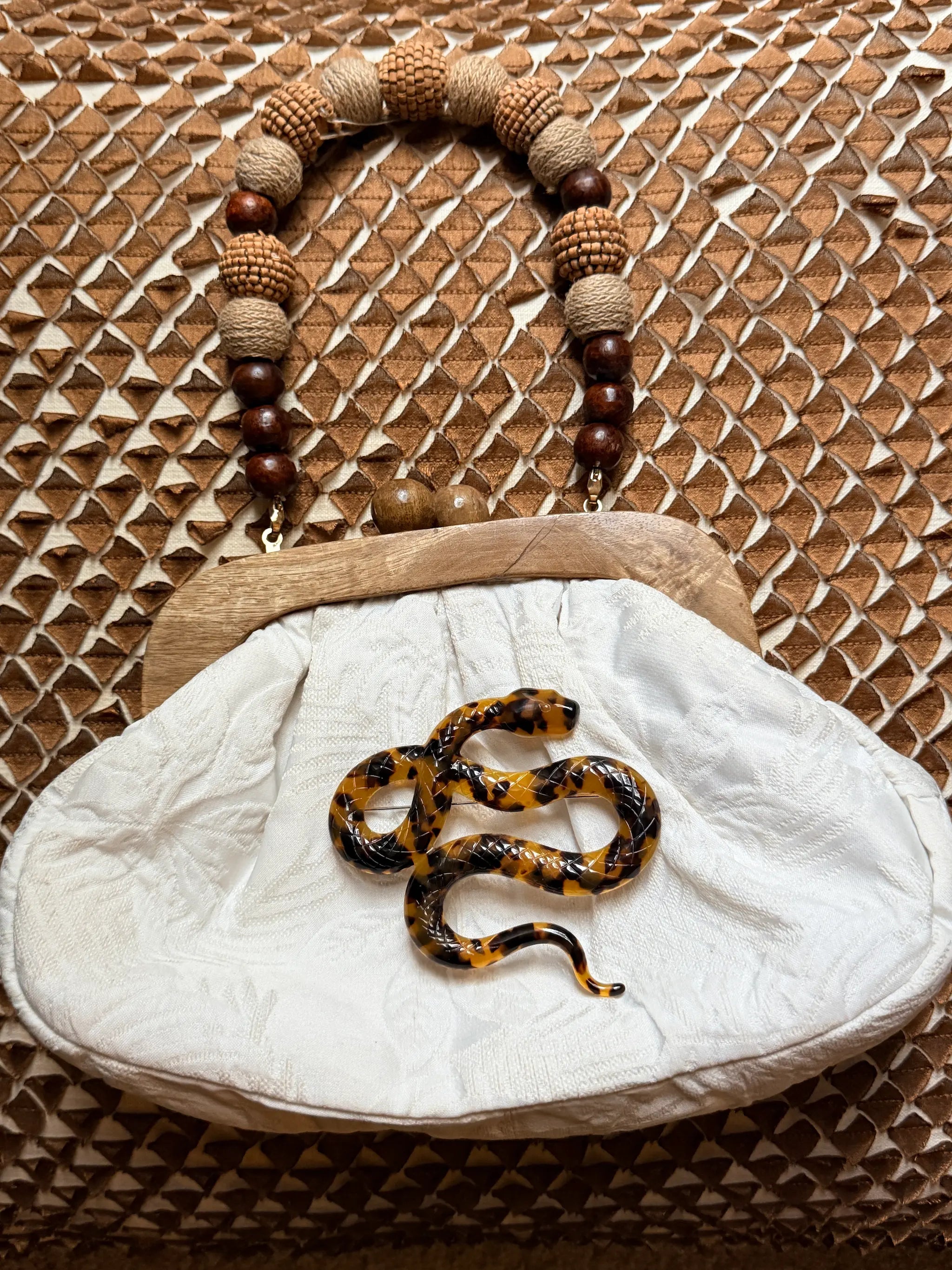 A vintage-style clutch handbag with a white fabric body, a light wood frame, and a beaded handle made of brown and beige beads. The bag is decorated with a realistic Broochella Animalia black and brown patterned snake brooch on the front, set against a brown woven background.
