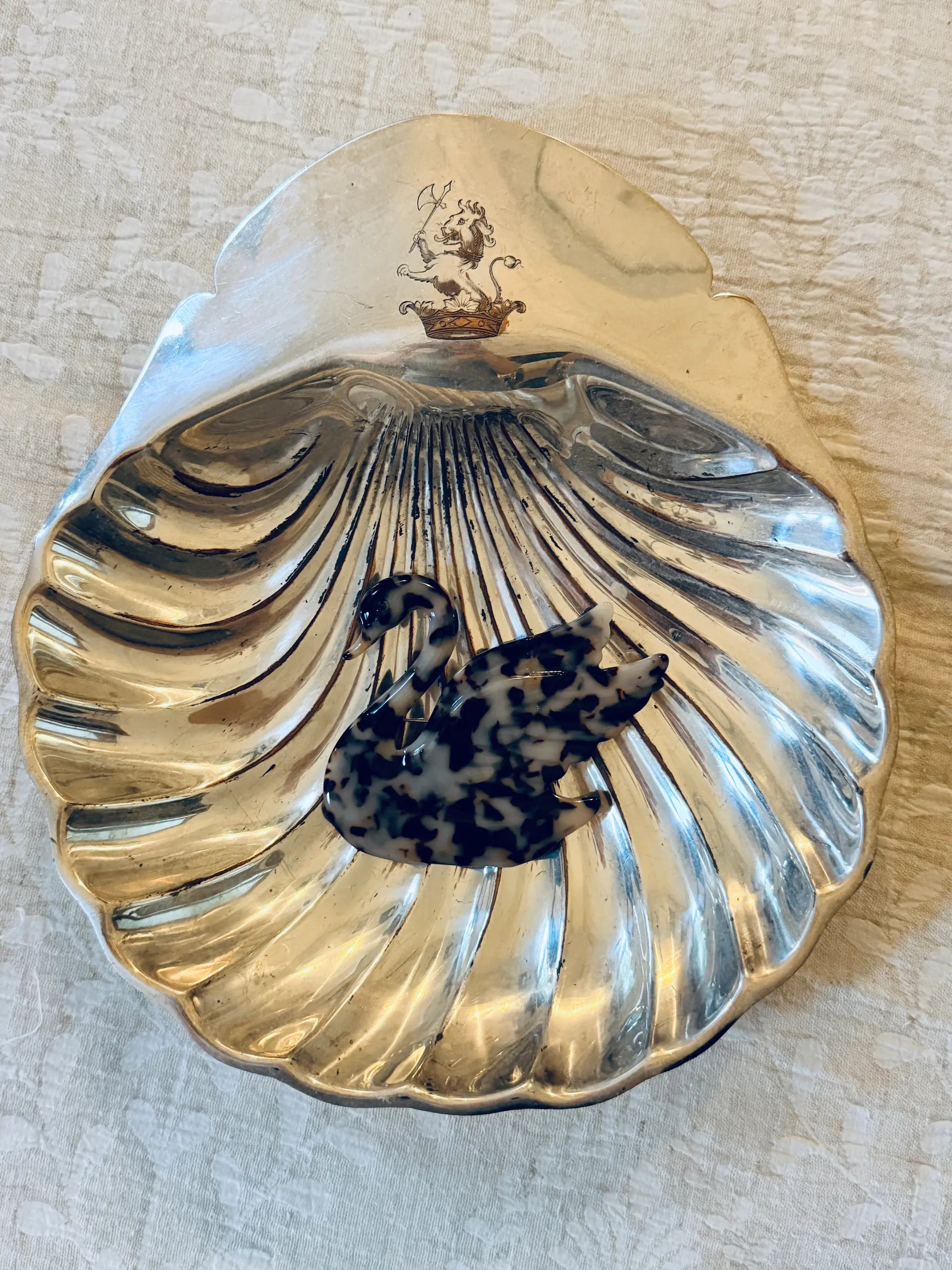 A silver-toned shell-shaped dish with a Broochella Animalia tortoiseshell swan brooch  placed inside it, photographed on a light textured fabric background.