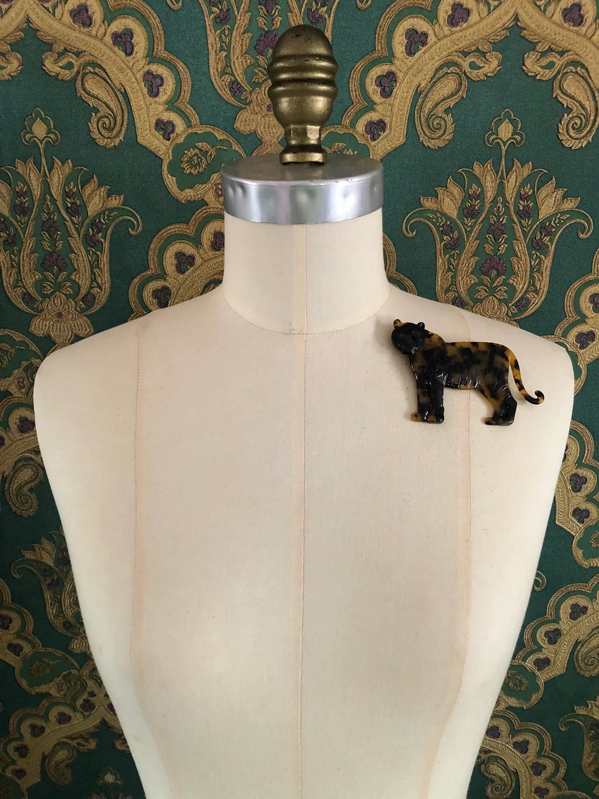 Broochella Animalia handmade tortoiseshell tiger brooch in Italian brown cellulose acetate. It is pinned to a mannequin The background is green and gold damask wallpaper. 