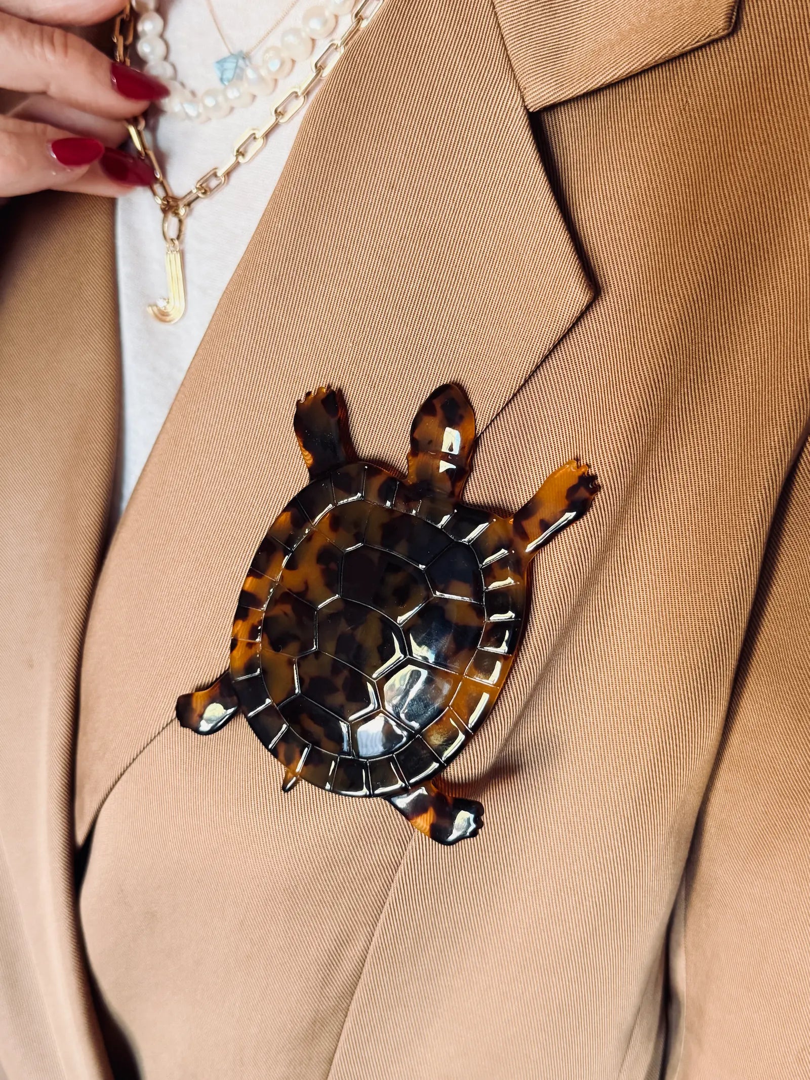 A Broochella Animalia collection tortoiseshell turtle-shaped brooch pinned to a tan blazer, with a person's hand and red nail polish visible at the top left.