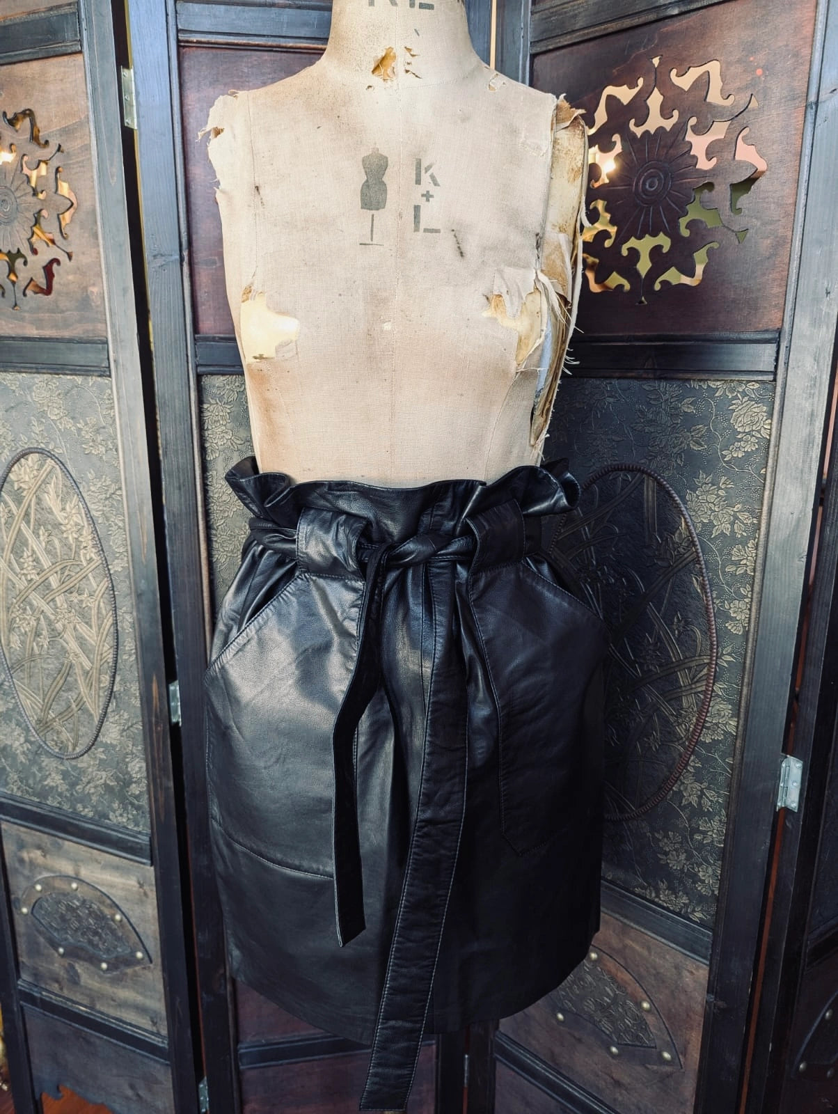 Broochella Anine Bing Black Leather Mini Skirt – Size Small. It has a paper bag waist and a tie up belt. It is pictured on a mannequin. The background is an ornate dark wood room divider
