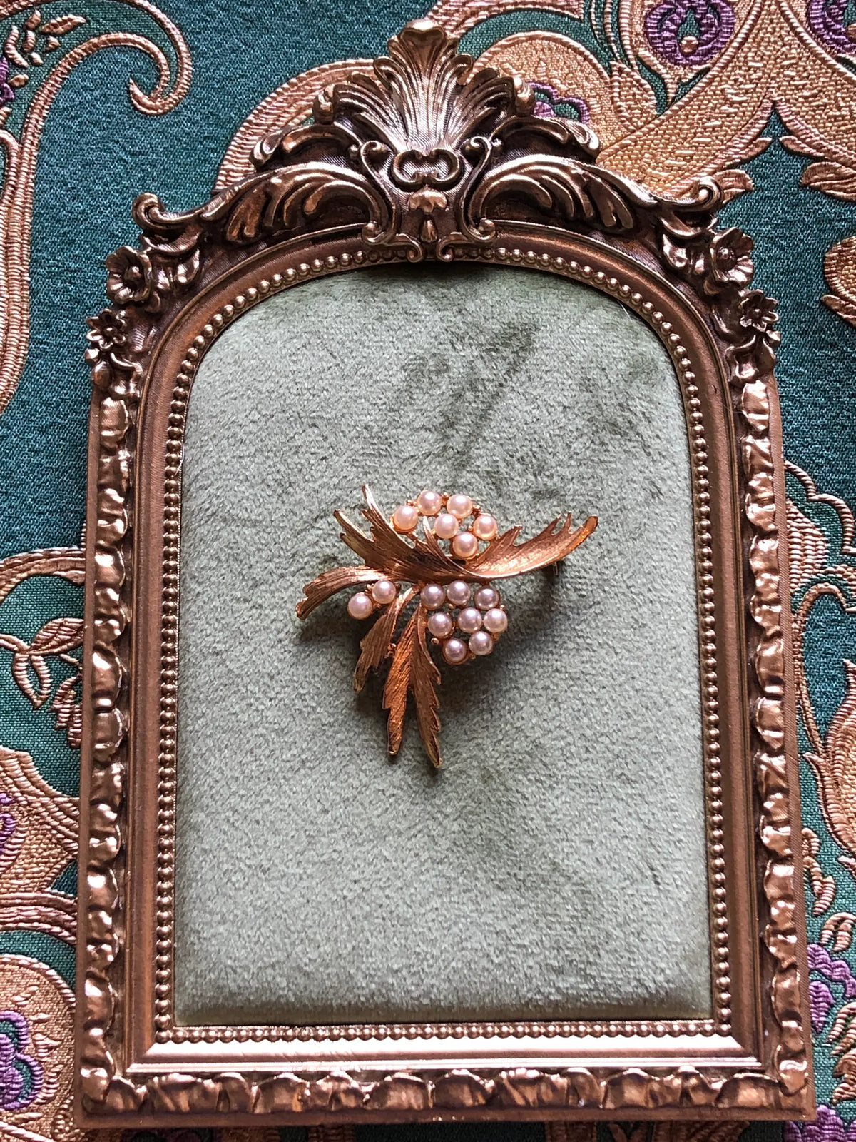 Broochella Antonella Vintage Pearl Leaf Brooch featuring clusters of ivory faux pearls amongst sculptural textured gold-tone leaves. It is presented on a green tray with an ornate gold frame. The background is green and gold damask wallpaper. 