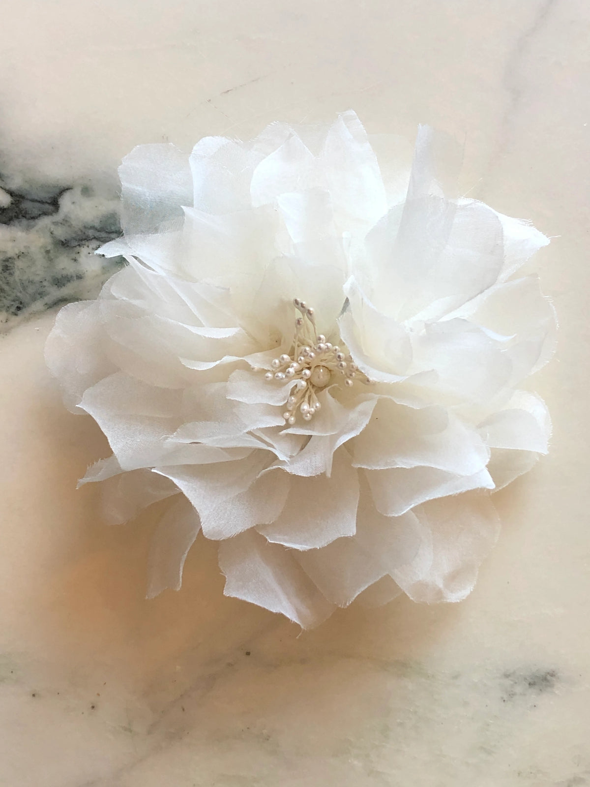 Broochella Aubrieta Bridal Flower Brooch in ivory, featuring soft layered fabric petals with delicate pearl detailing at the centre, photographed on a pale cream marble background with soft grey-green veining.
