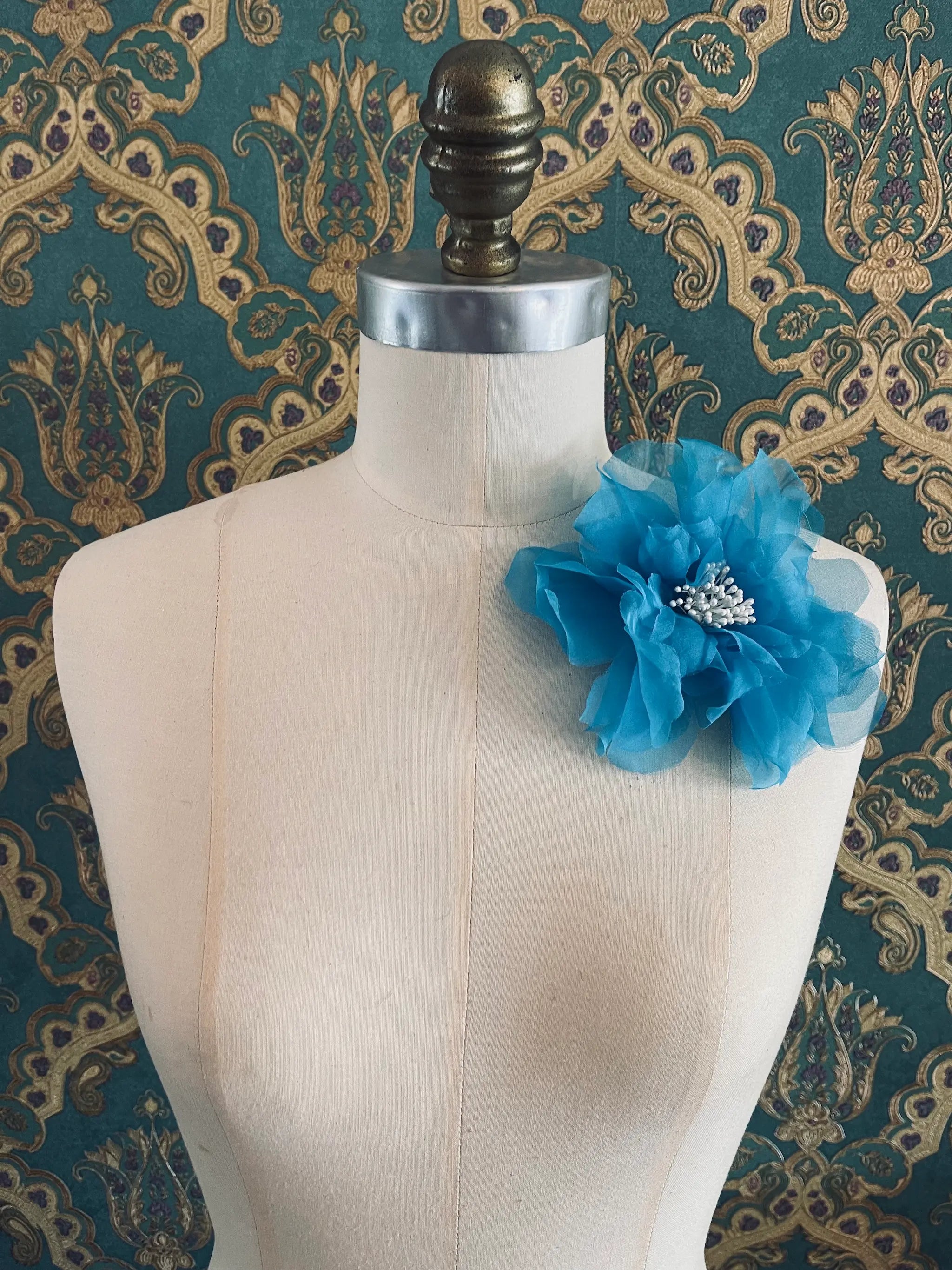 Broochella Aubrieta handmade fabric flower brooch in blue. Pictured on a mannequin. The background is green and gold damask wallpaper