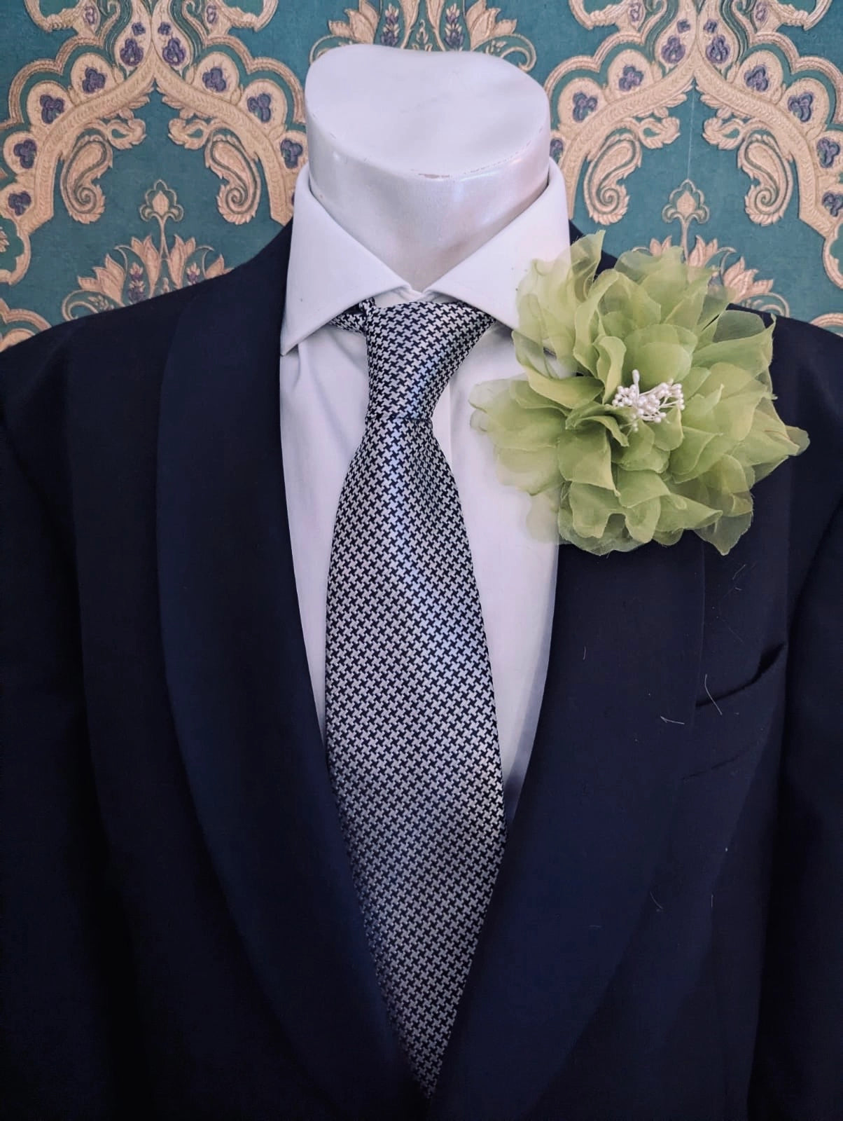 A black suit jacket displayed on a white mannequin torso, styled with a white dress shirt, a patterned grey and white tie, and a Broochella men's chartreuse  Aubrieta flower brooch against a teal and gold damask wallpaper background.