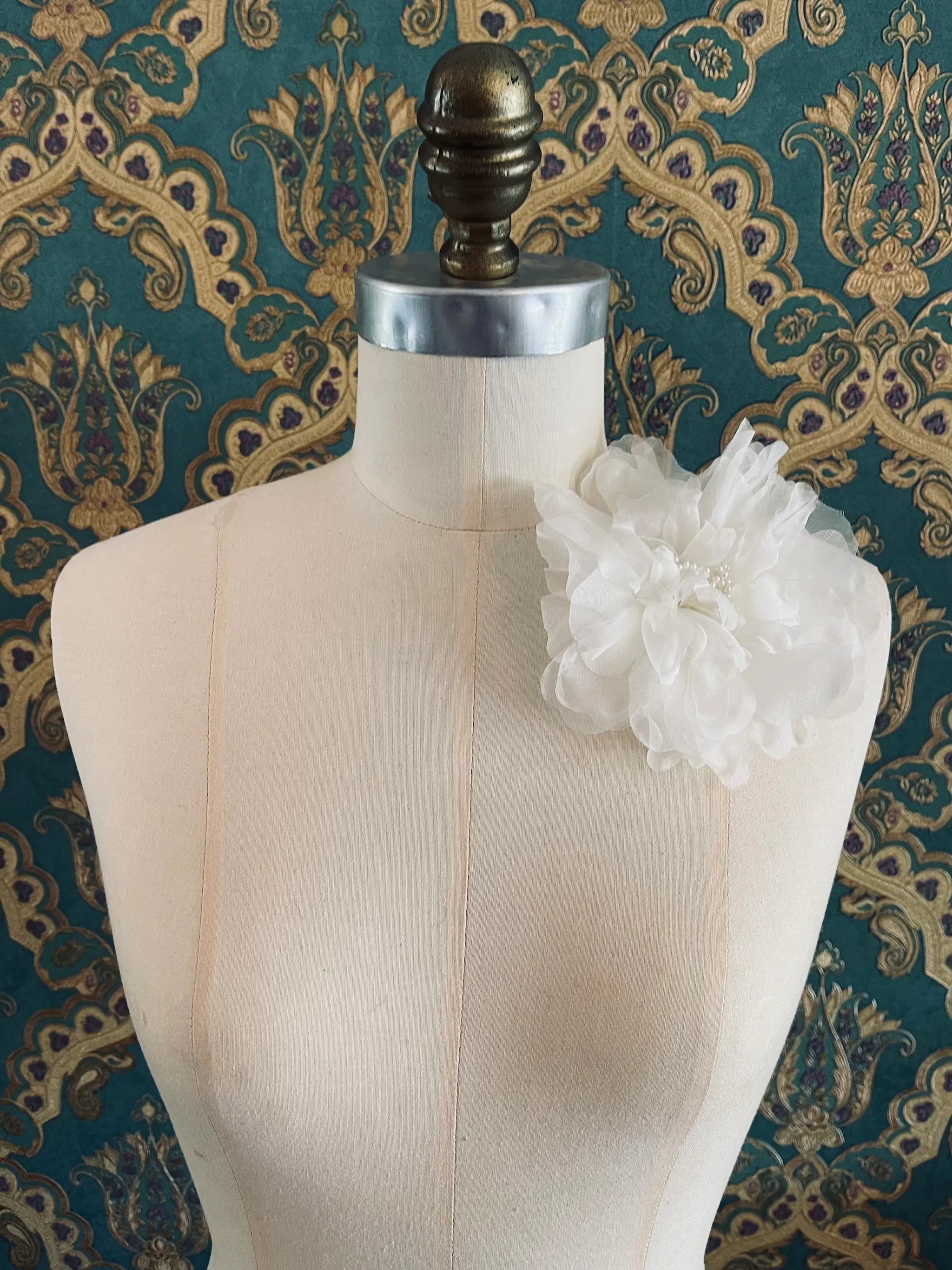 Broochella Aubrieta handmade fabric flower brooch in ivory. Pictured on a mannequin. The background is green and gold damask wallpaper