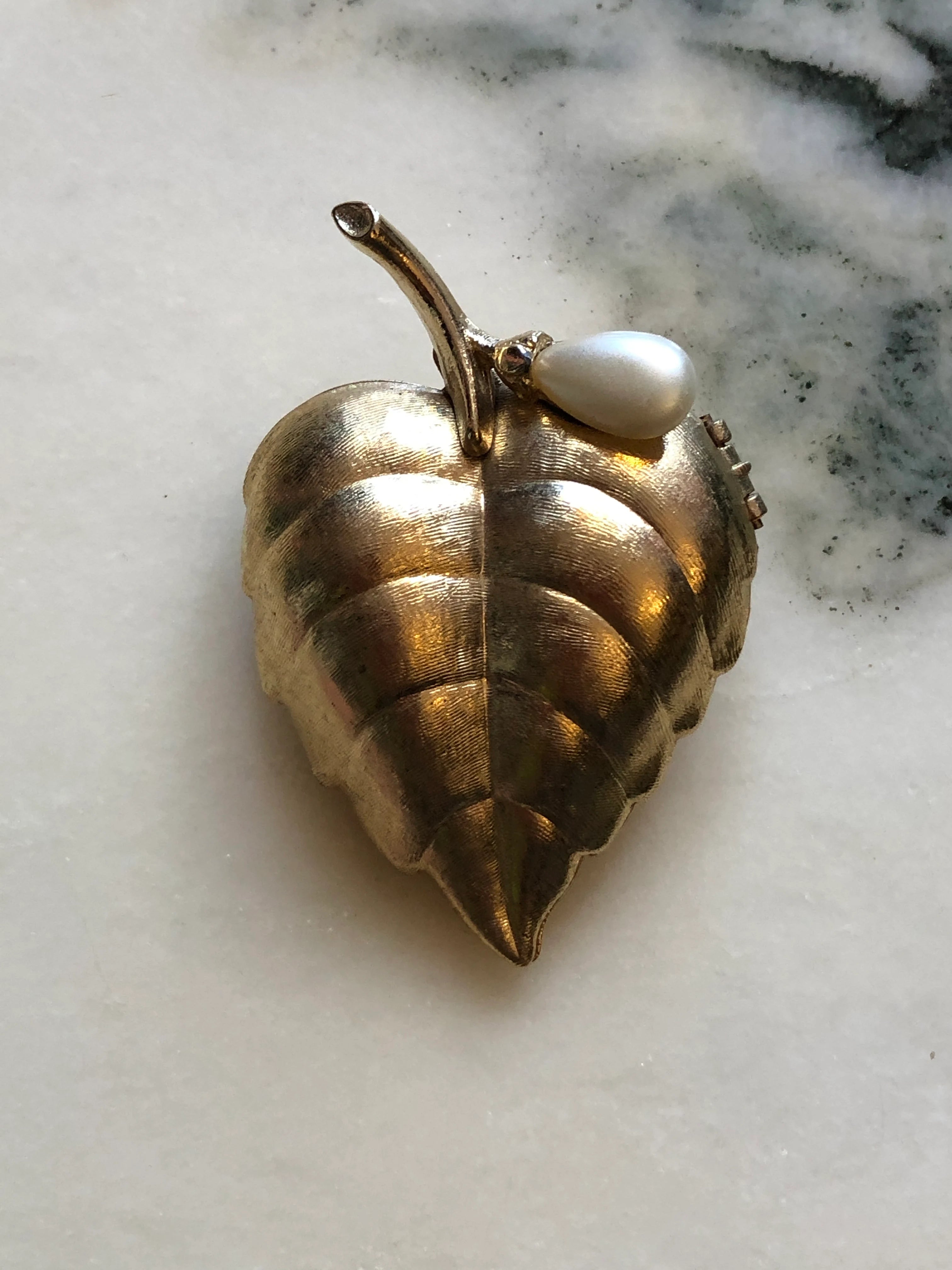 Broochella Avon vintage gold-tone leaf perfume glacé brooch with engraved detailing and a faux pearl accent. The background is white marble.