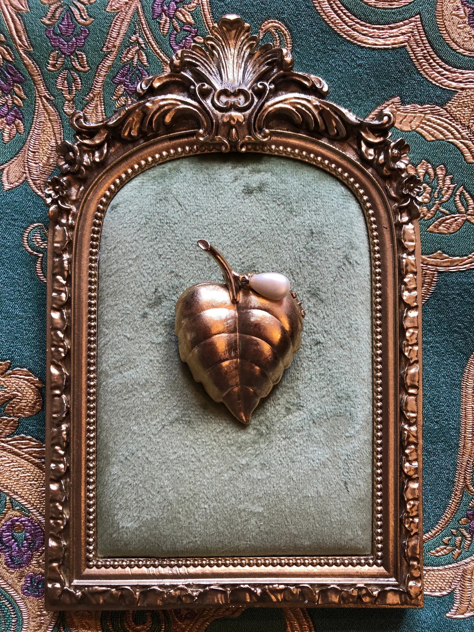 An ornate antique-style bronze frame with a carved floral crest and beaded border, enclosing a textured sage green fabric insert with a Broochella Avon Golden Leaf perfume brooch in the centre. It is set against a patterned teal and gold brocade background.