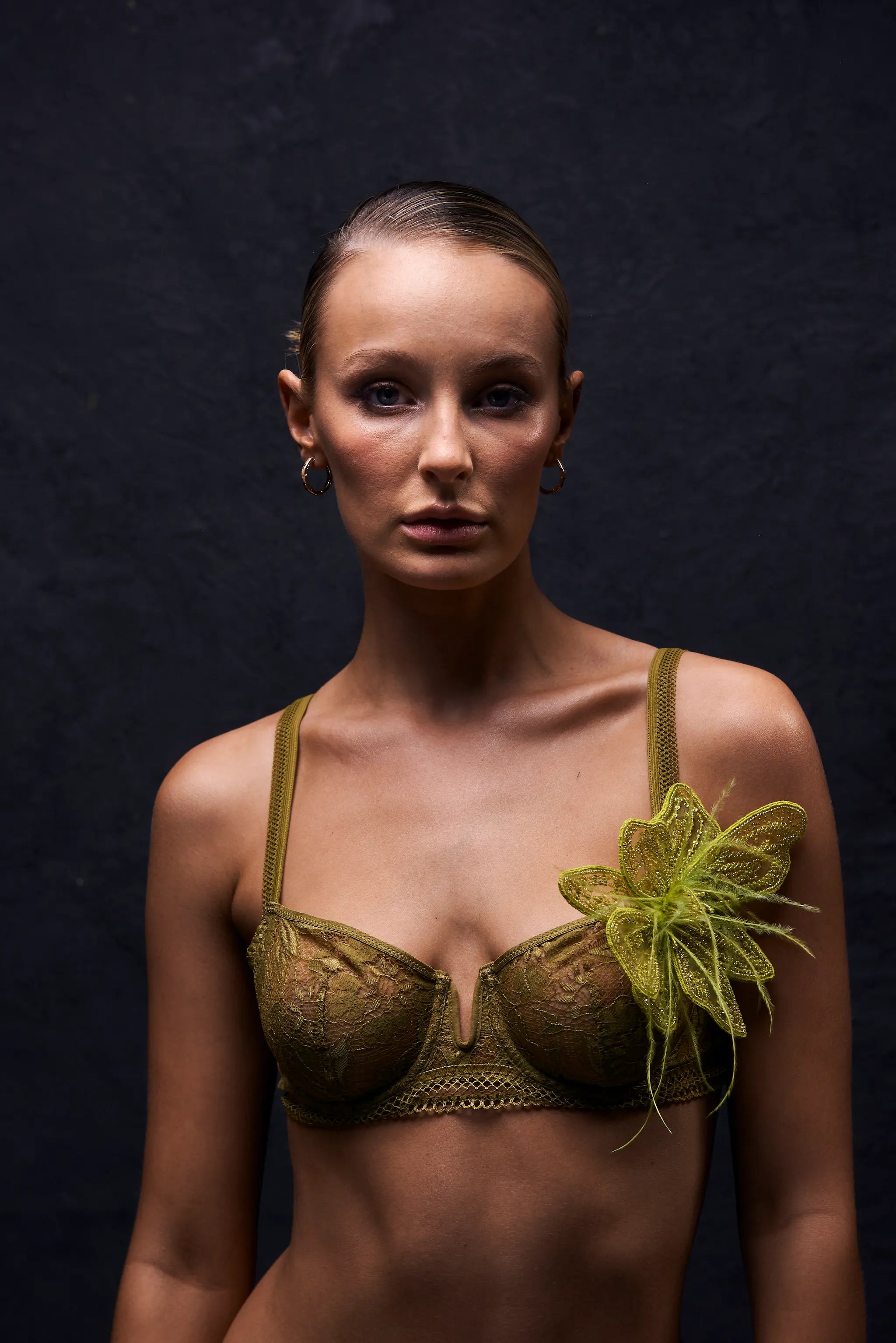 A model wearing an olive green lace bra with a Broochella azalea chartreuse  textured green flower brooch on the left cup, set against a dark textured studio background.