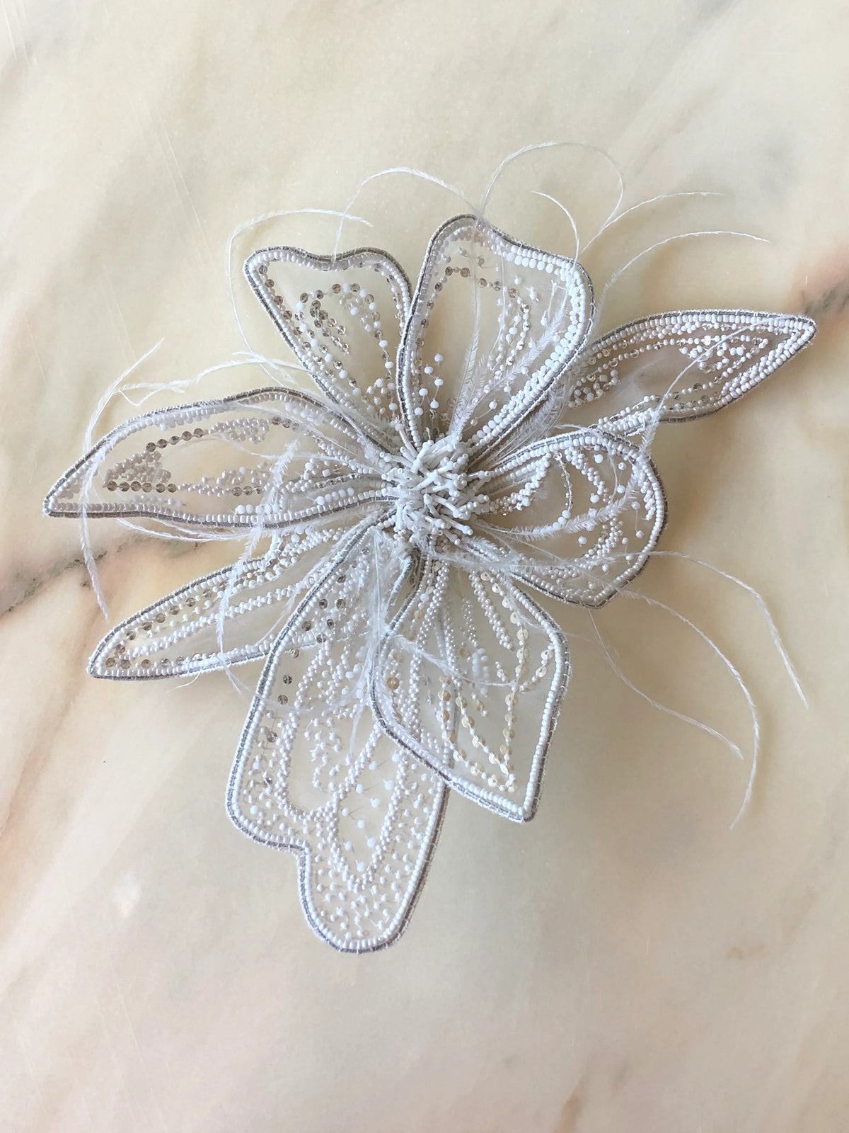 Broochella Azalea Flower Bridal Brooch Large in white, featuring sculptural beaded and sequinned petals with delicate white feather accents, photographed on a cream marble background.