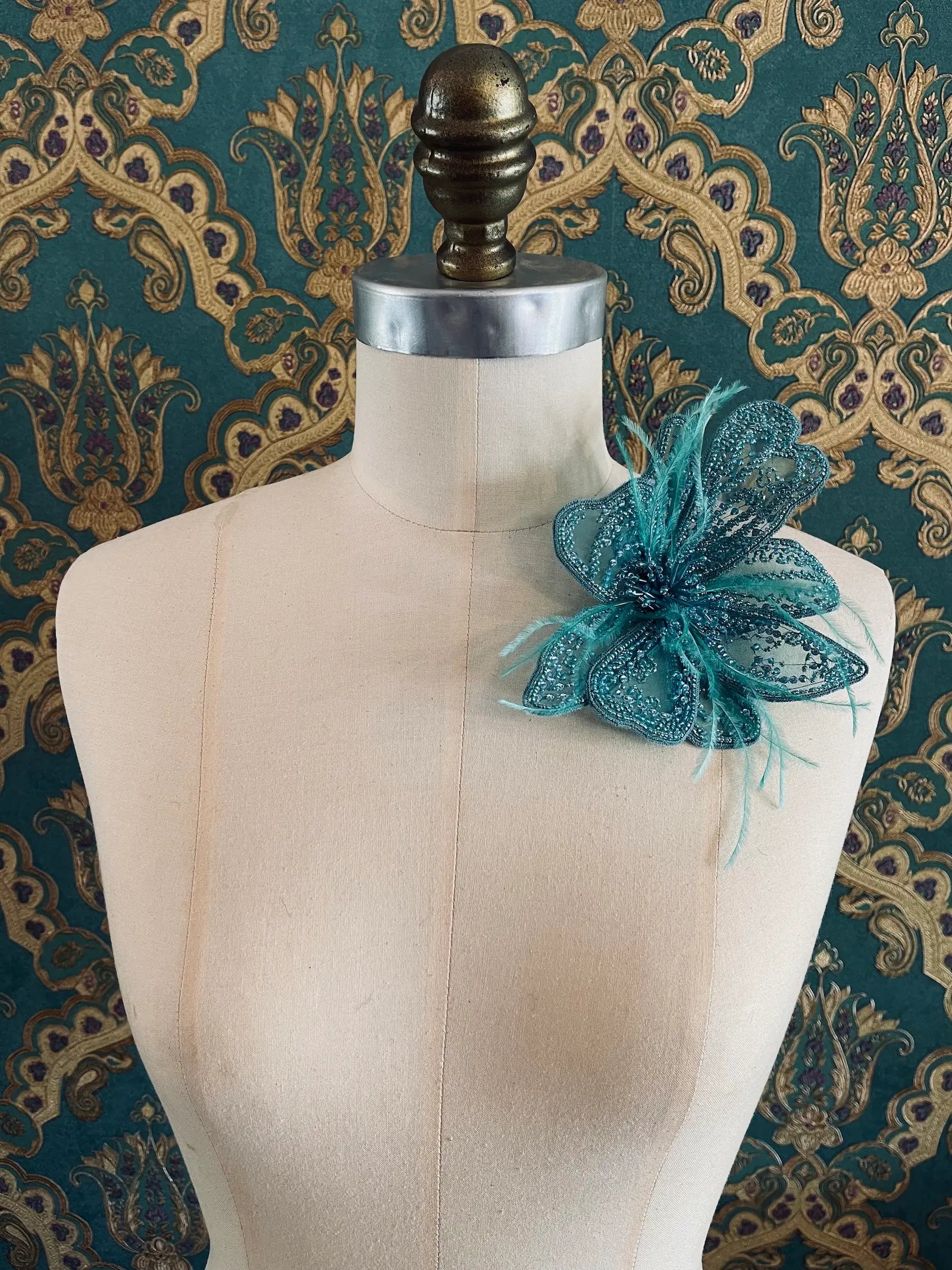 Broochella Azalea hand beaded flower brooch in blue. Pictured on a mannequin. The background is green and gold damask wallpaper
