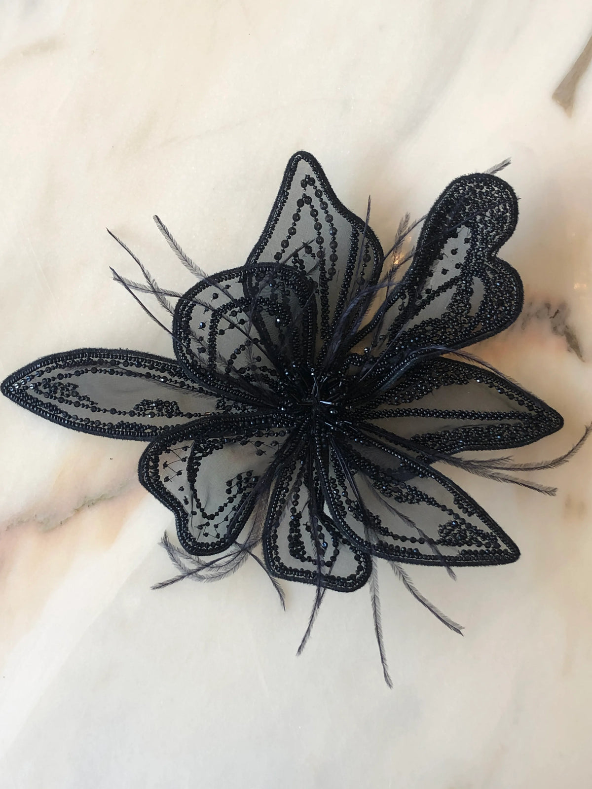 Broochella Azalea Flower Hand-Beaded Bridal Brooch in black, large size, featuring layered sheer petals embellished with black beads, sequins and delicate feathers, photographed on a white marble background with soft grey-green veining.