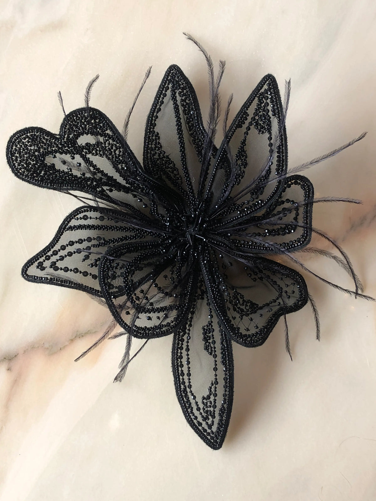 Broochella Azalea Large handmade designer flower brooch in sheer black organza, intricately hand-beaded with black sequins and beads and finished with delicate black feather details, photographed on cream marble with natural grey veining.
