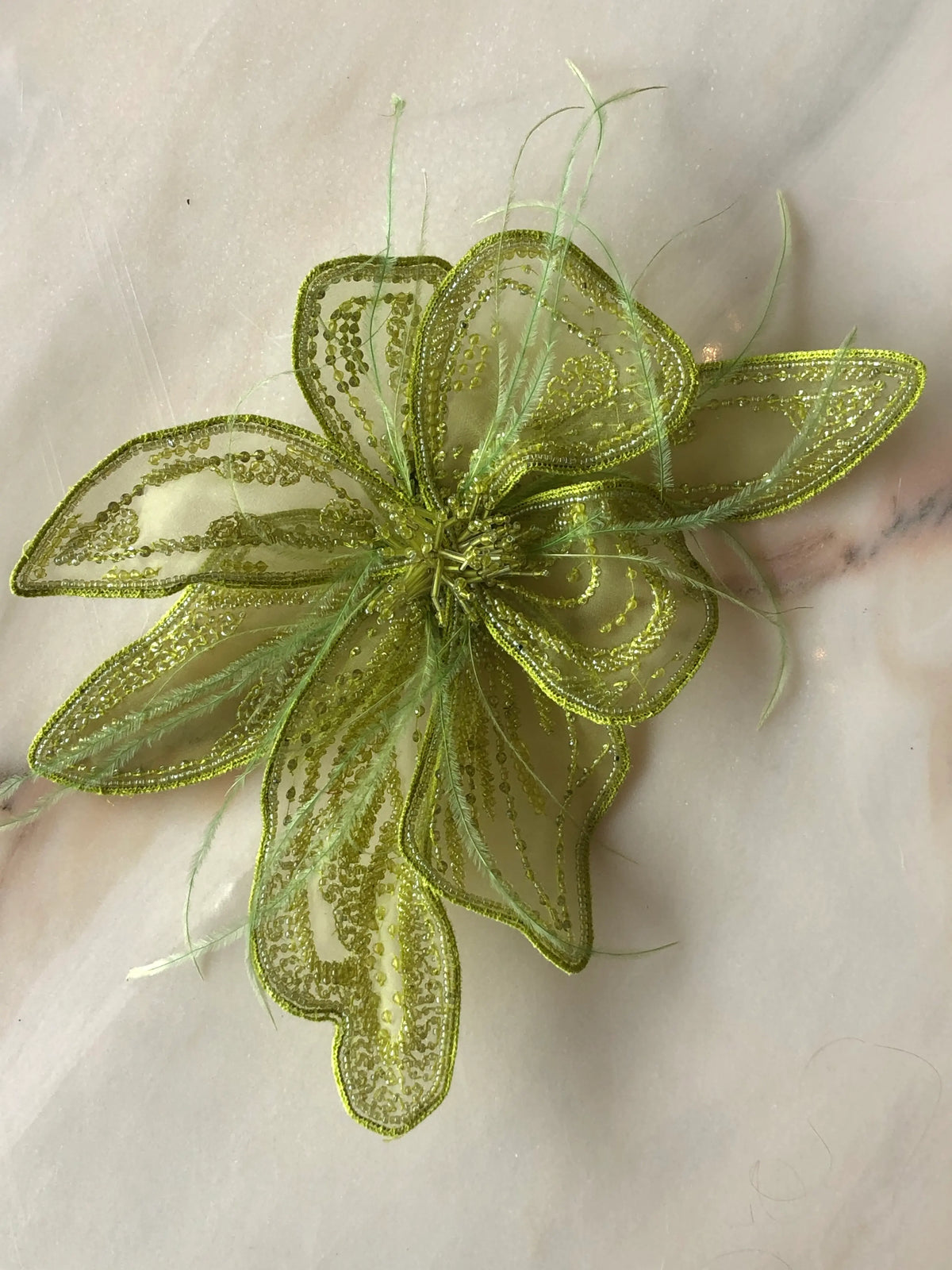 Broochella Azalea Large handmade designer flower brooch in sheer chartreuse organza, intricately hand-beaded with tonal green sequins and beads and finished with delicate feather details, photographed on cream marble with natural grey veining.