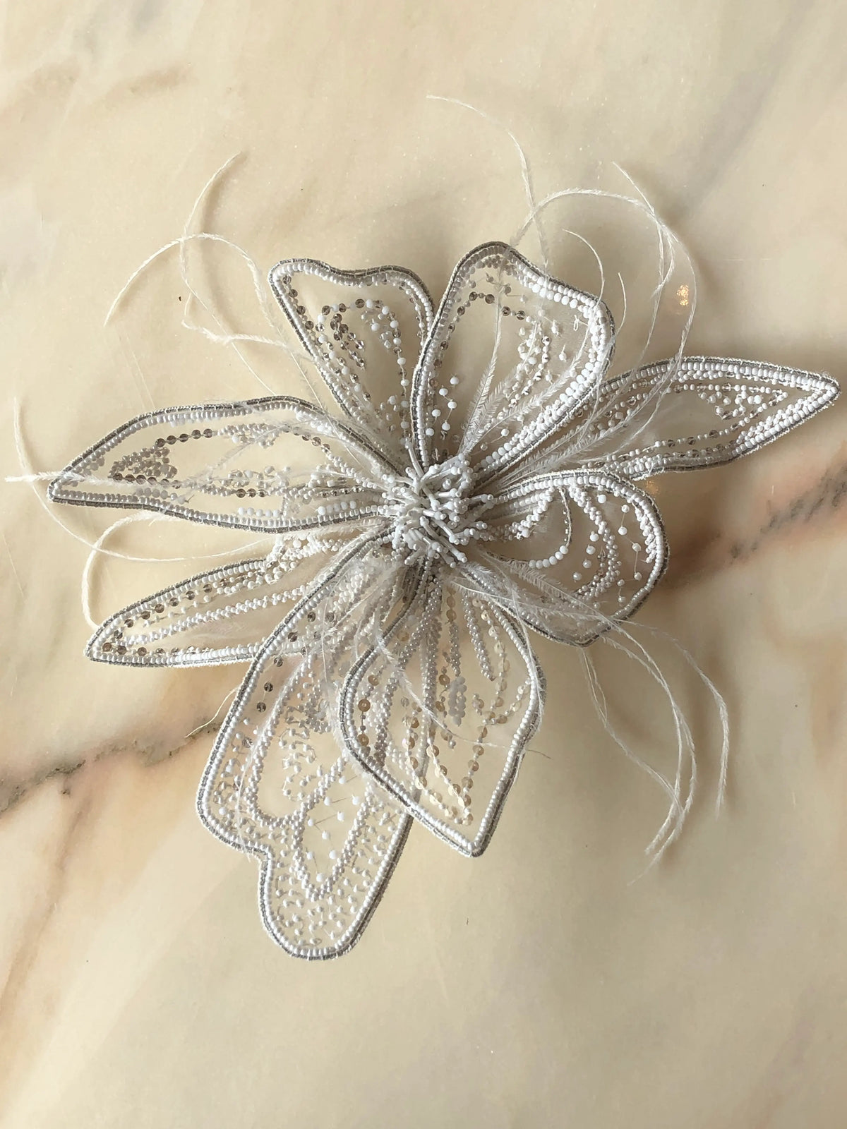 Broochella Azalea Large handmade designer flower brooch in sheer white organza, intricately hand-beaded with white and silver sequins and finished with delicate white feather details, photographed on cream marble with natural grey veining.
