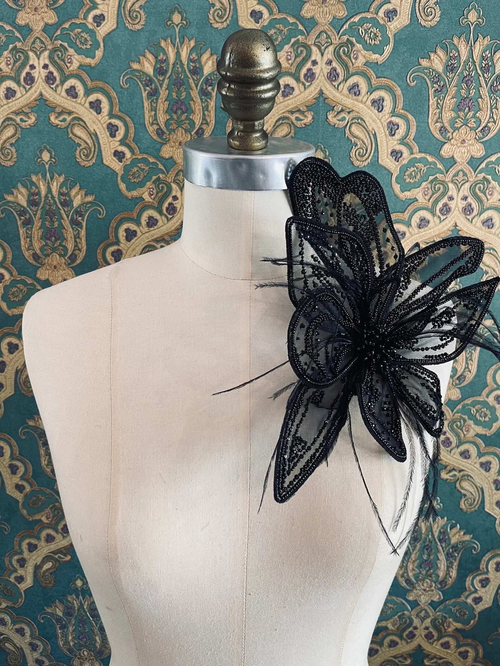 Broochella Azalea large handmade hand beaded flower brooch in black. The brooch is pinned to a mannequin. The background is green and gold damask wallpaper.