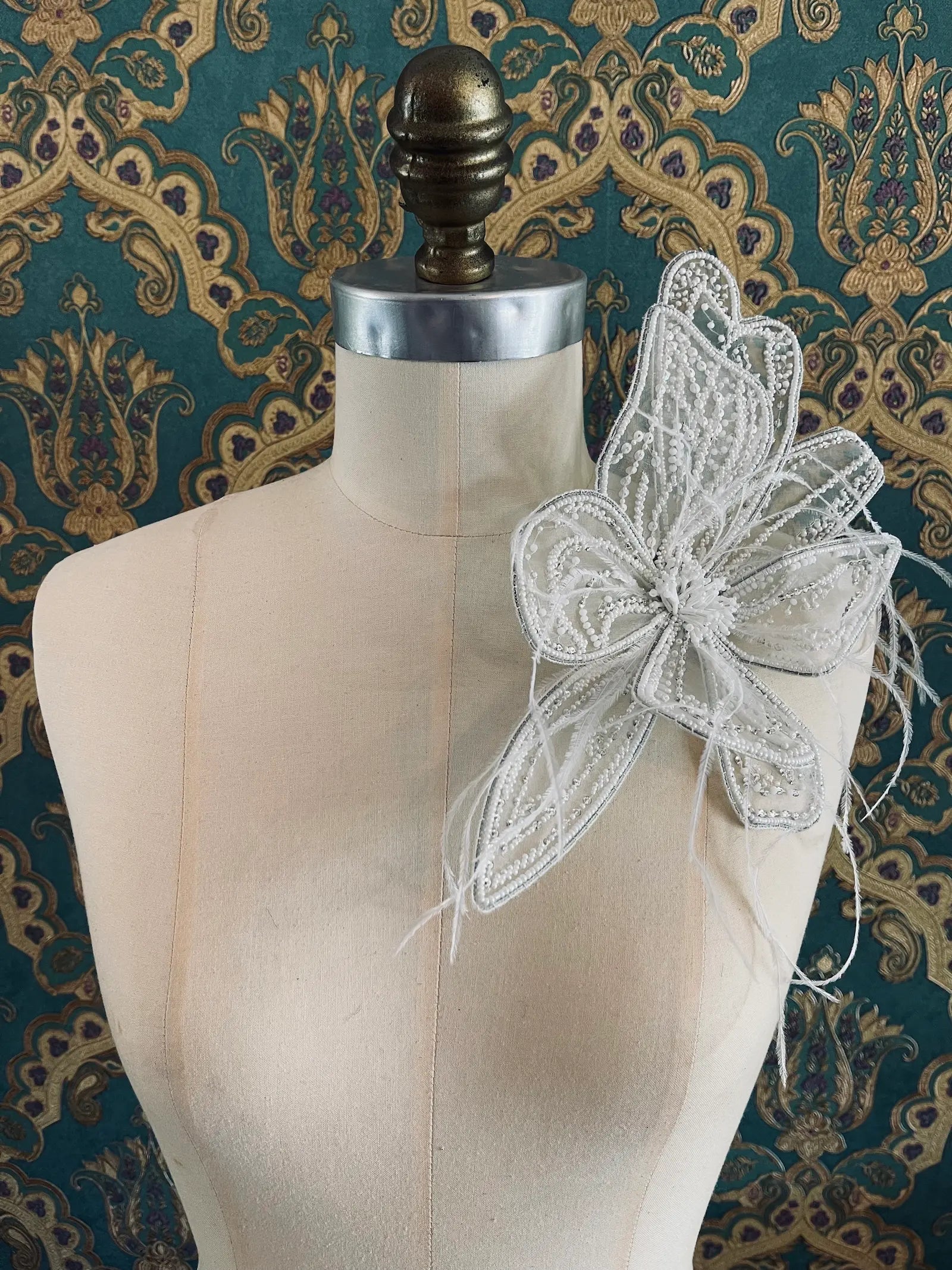 Broochella Azalea large handmade hand beaded flower brooch in white. The brooch is pinned to a mannequin. The background is green and gold damask wallpaper.