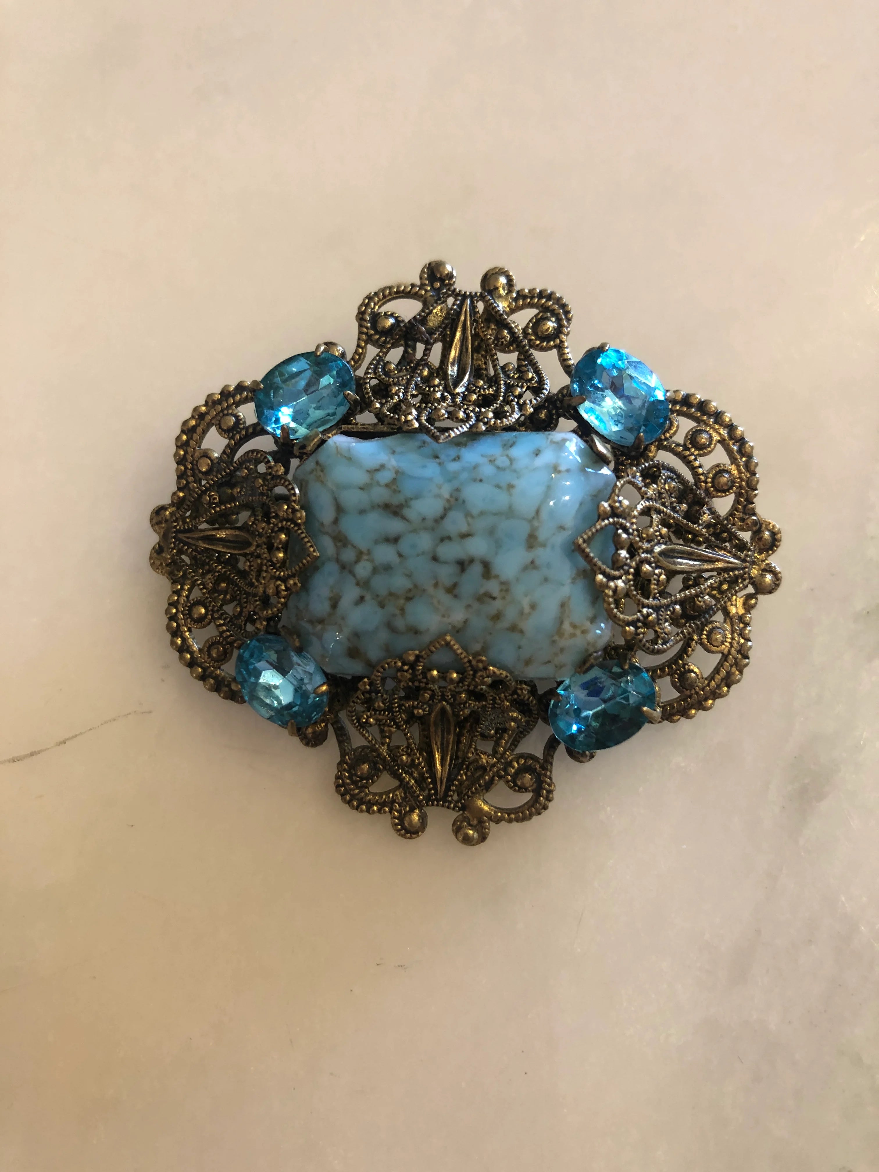A vintage Czech Azzurra gold-tone filigree brooch featuring a large square turquoise marbled cabochon centre stone set within an ornate granulated and scrollwork openwork frame, accented with six faceted aqua blue crystal rhinestones at the cardinal points. Pictured on a white marble surface.