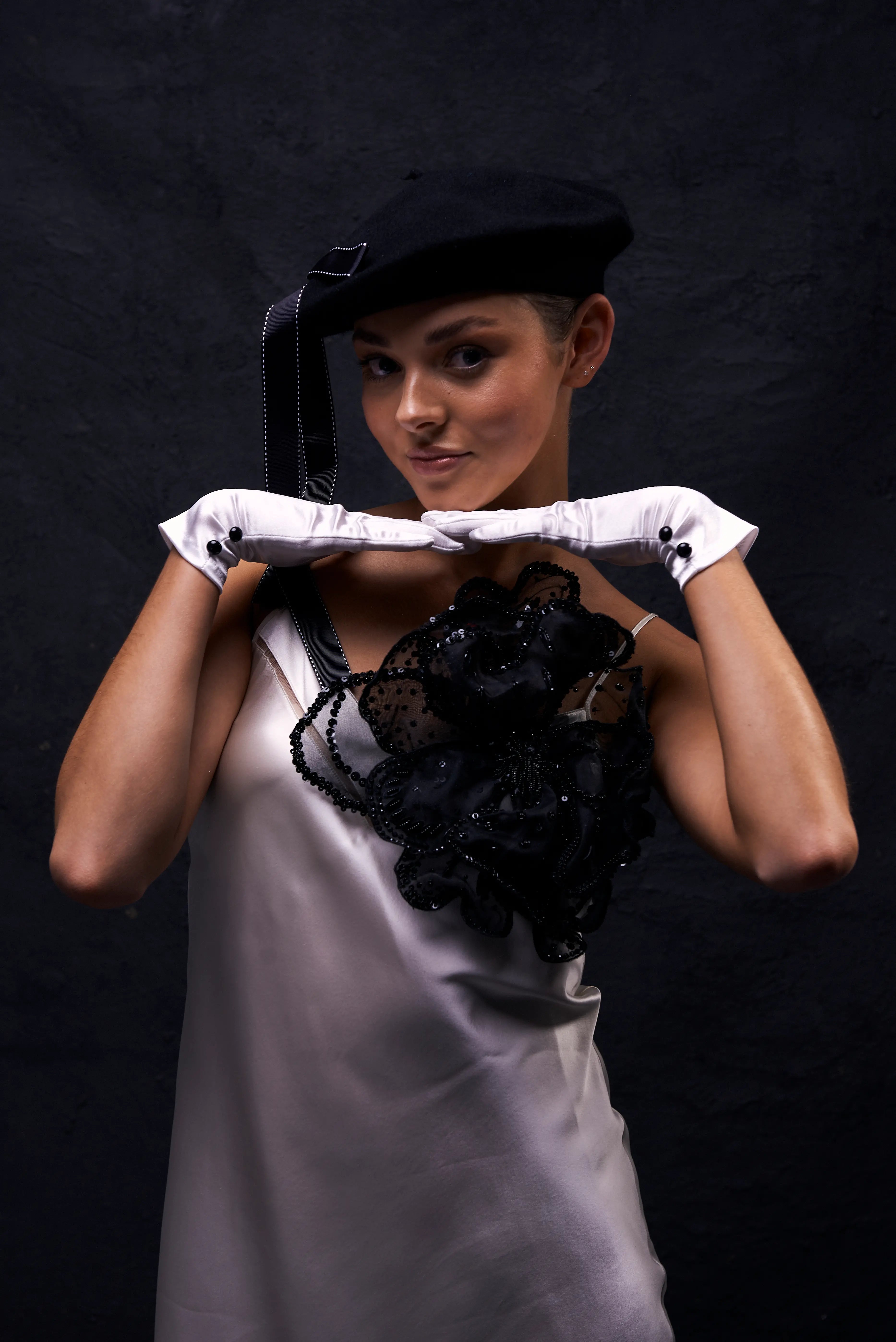 A woman wearing a black beret with a black ribbon, white gloves, and a light gray satin dress with a Broochella Baccara  large black lace floral brooch, posing against a dark textured background.