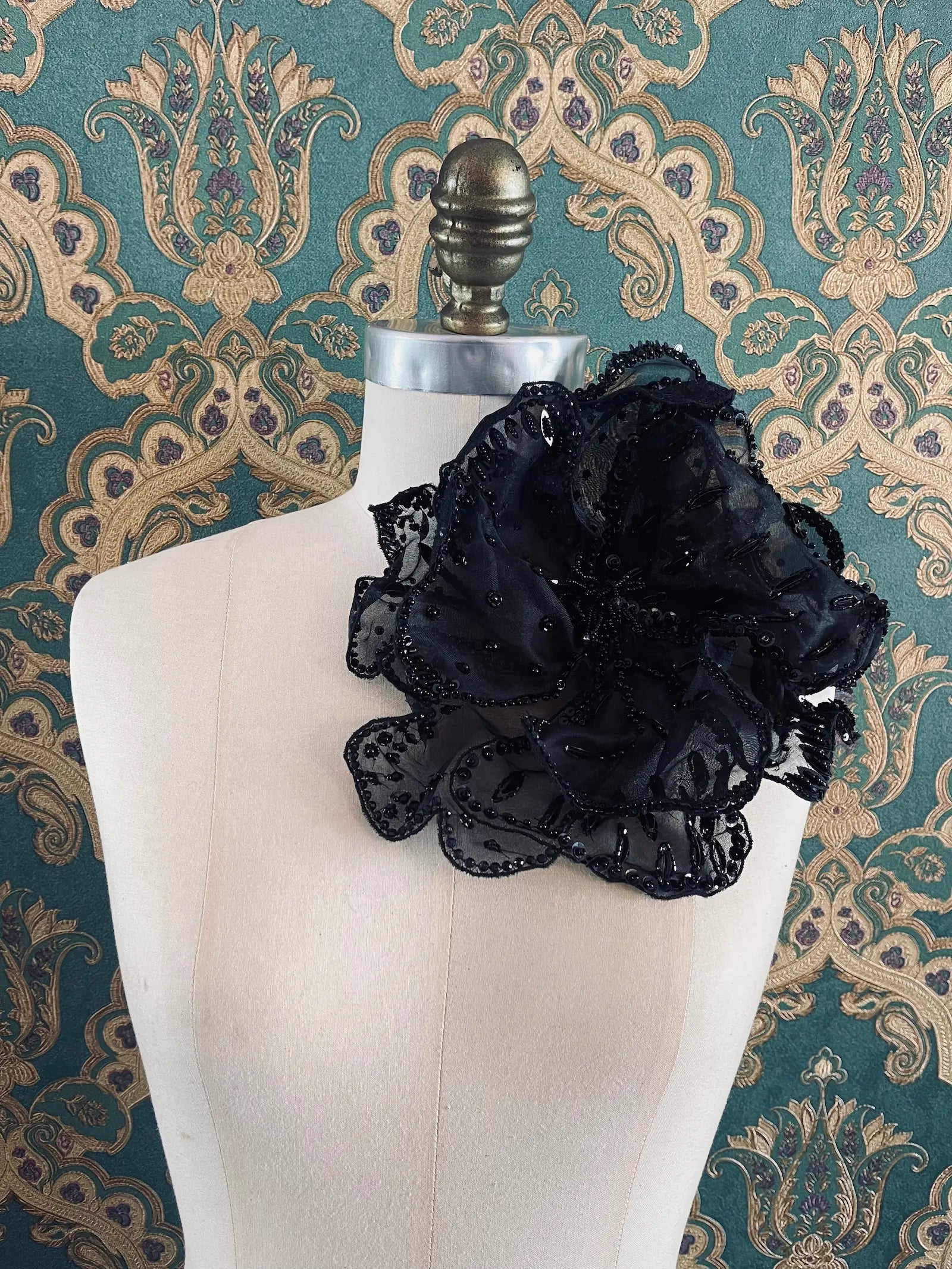 Broochella Baccara large black hand beaded flower brooch. Pictured on a mannequin. The background is green and gold damask wallpaper. 