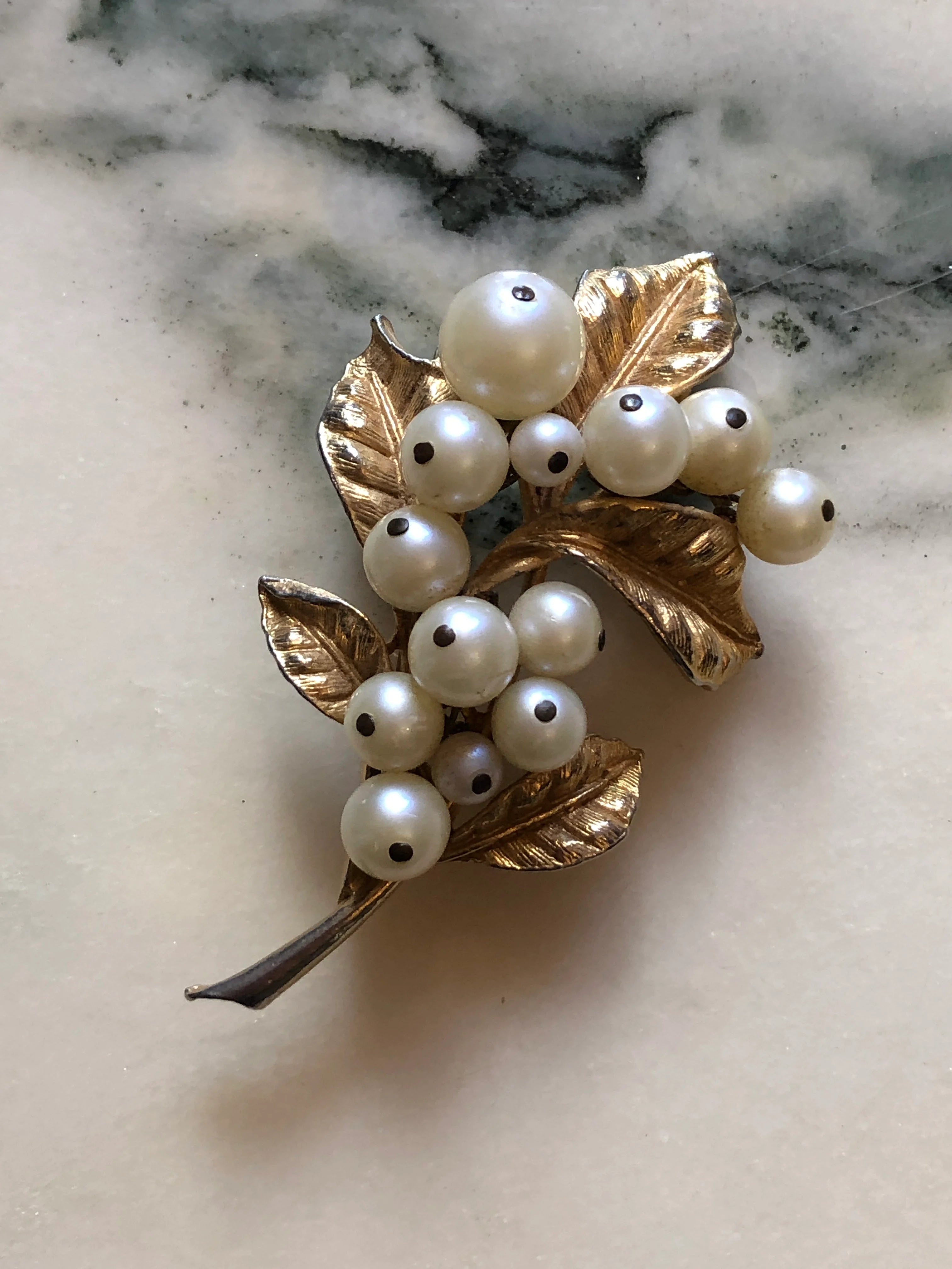 Broochella Bacche di Perla vintage gold-tone botanical brooch with textured leaves and graduated faux pearl clusters. Pictured on a white marble surface.