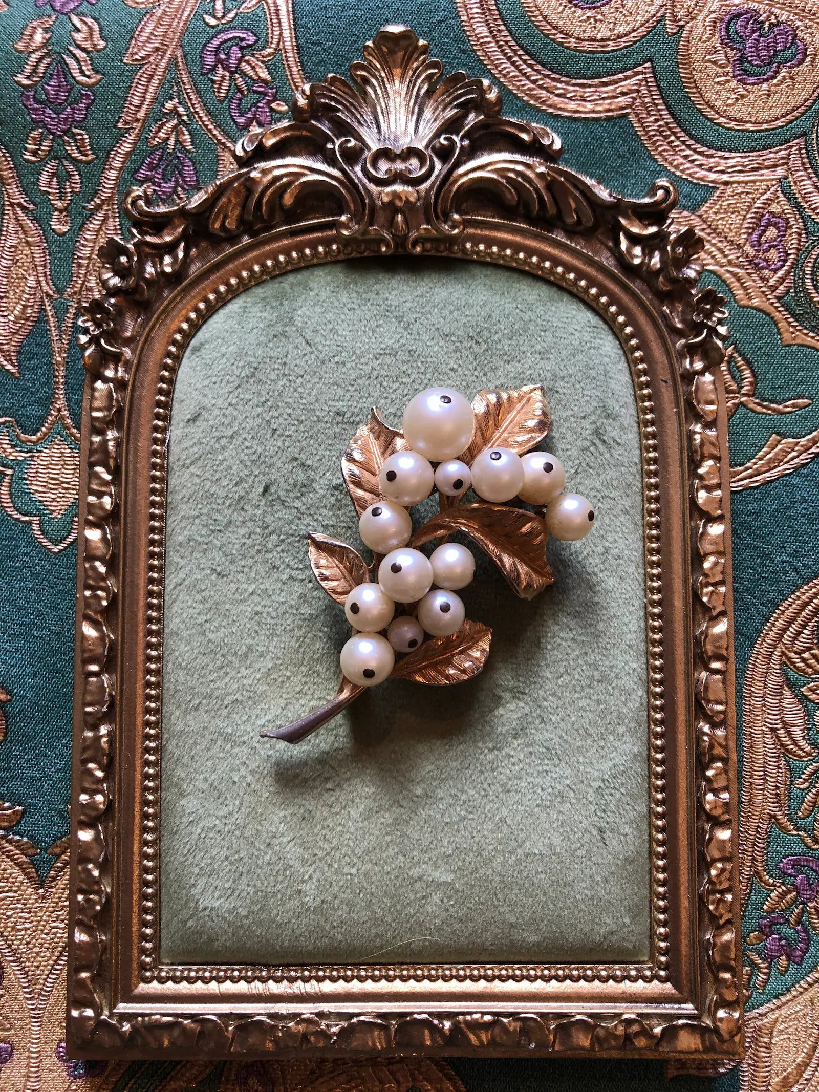 A Broochella Bacche di Perla vintage brooch featuring white pearl-like berries and metallic gold leaves, displayed on a green velvet surface inside an ornate gold frame.