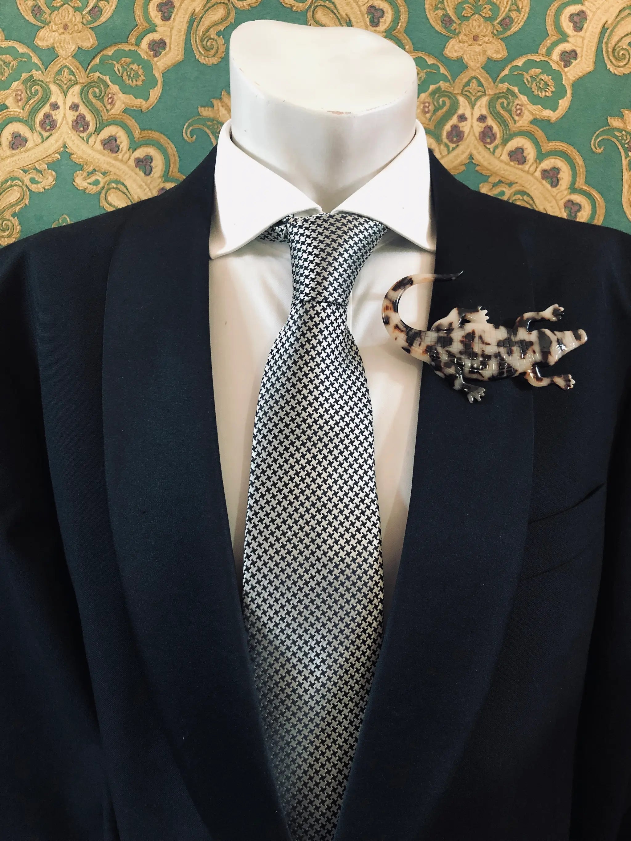 Broochella Men's beige Animalia tortoiseshell crocodile shaped brooch. It is made from Premium Italian cellulose tortoiseshell acetate. It is presented on a men's mannequin that is dressed in a black suit. The background is green and gold damask wallpaper. 